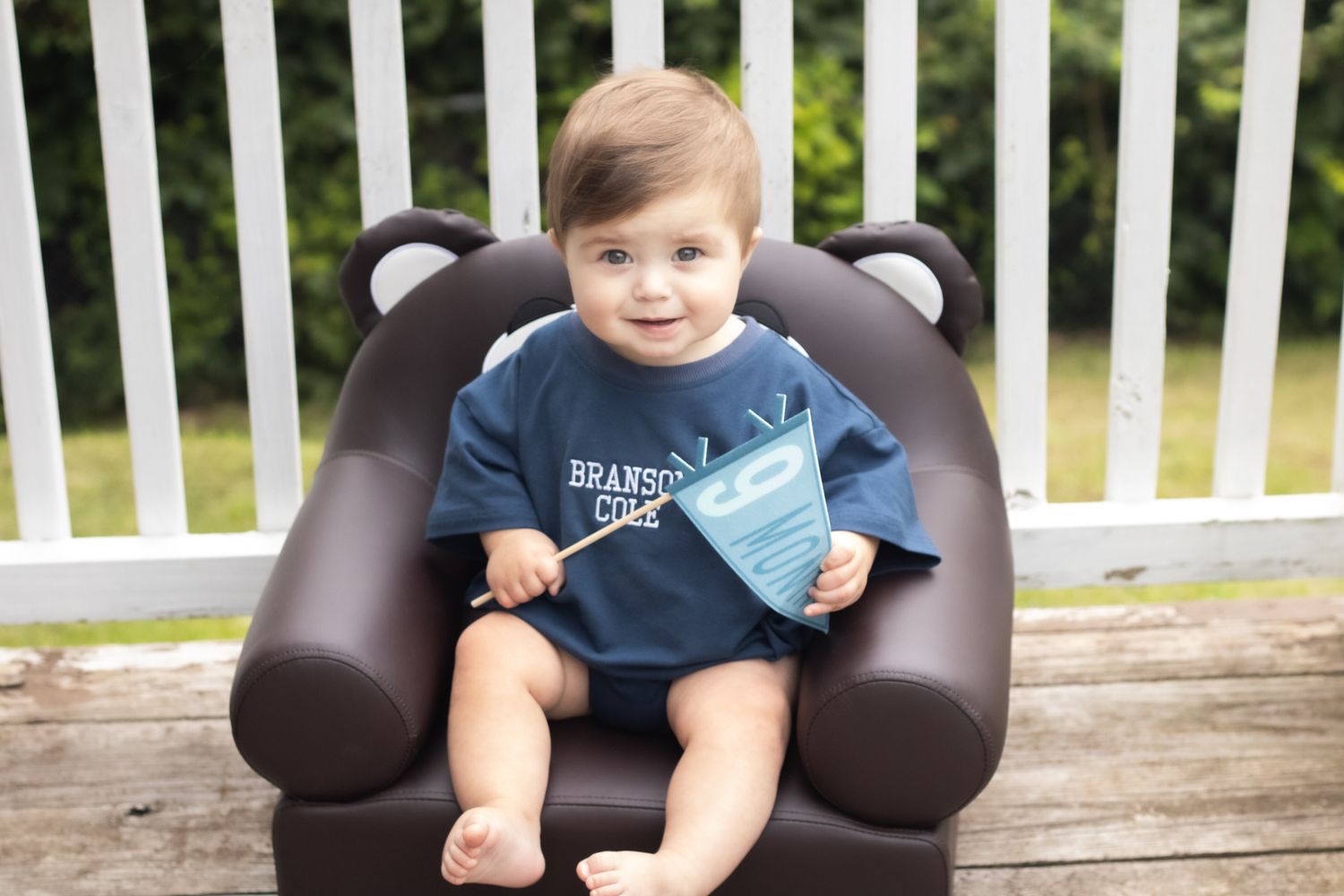 Cute baby boy sitting in bear chair outdoors holding a book on a wooden deck.