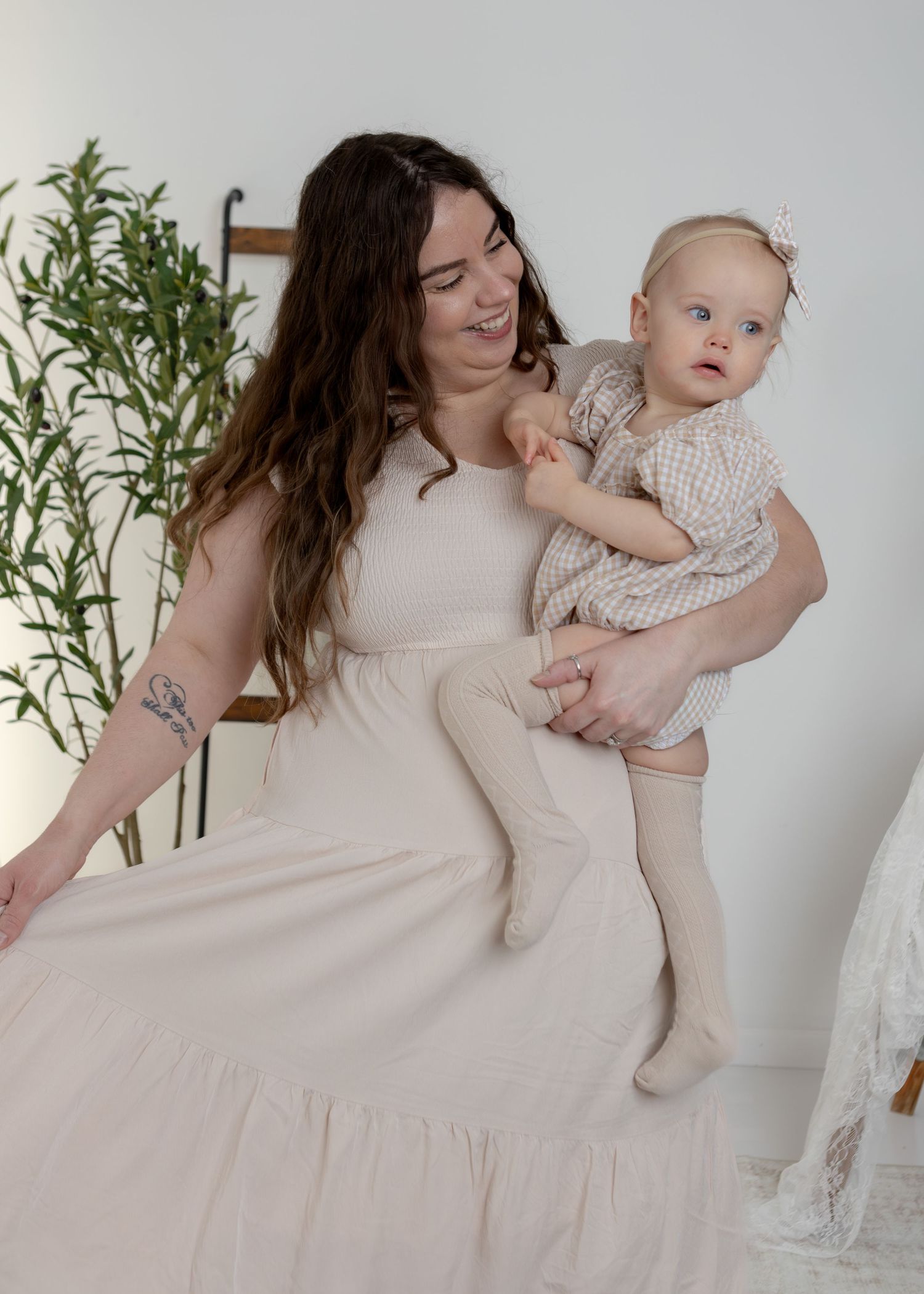 Smiling brunette mother in beige dress holding her blue-eyed baby girl in a bright studio.