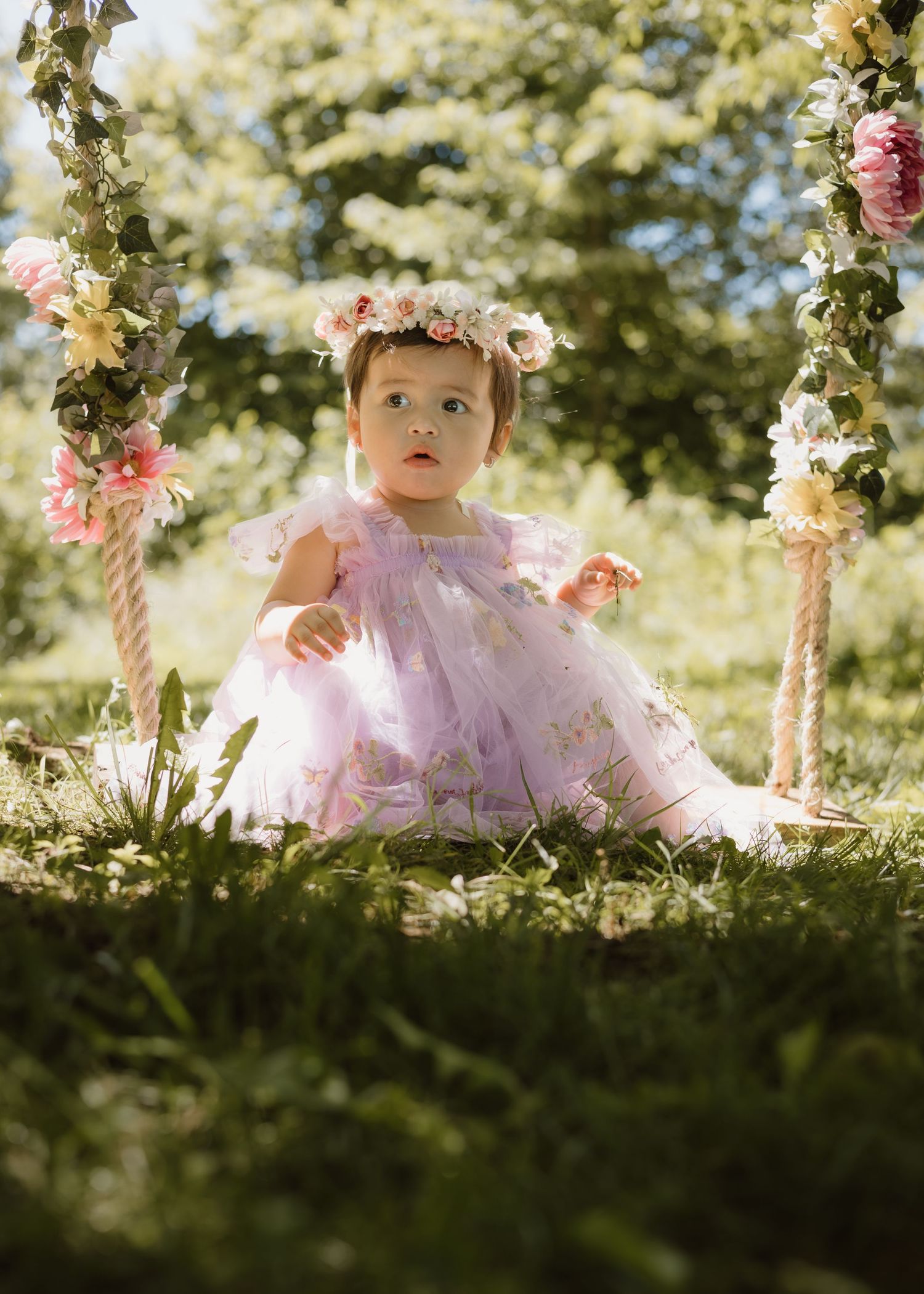 Sweet baby girl in lavender tulle dress and flower crown sitting on garden swing.