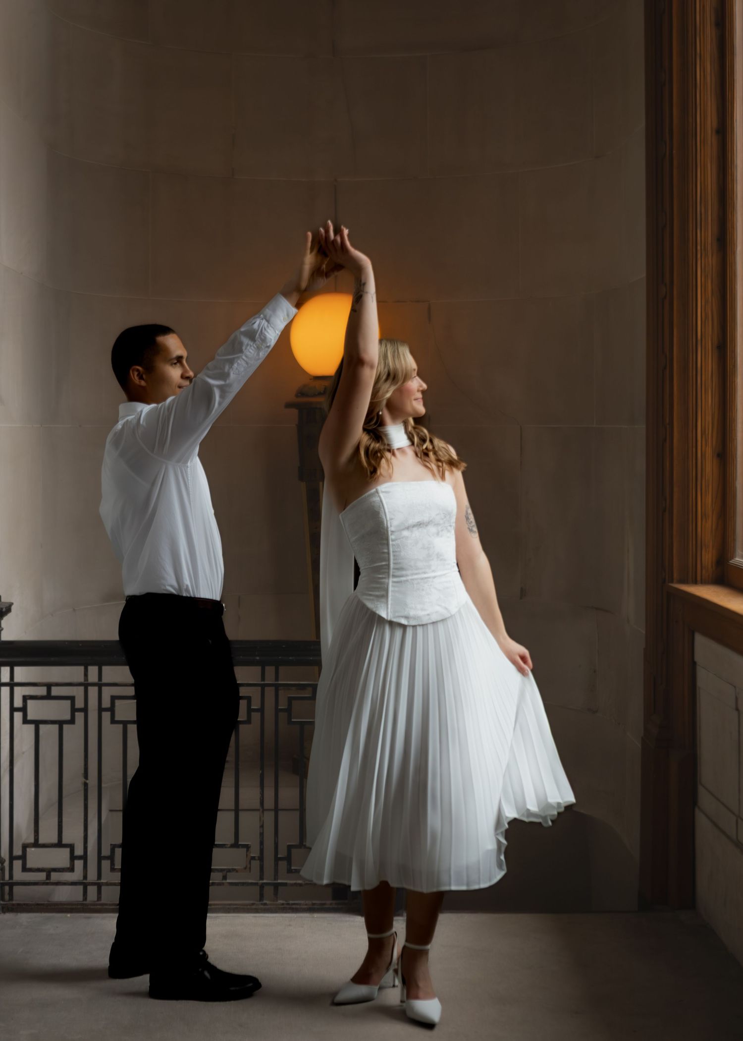 Man spinning woman in white dress near a glowing lamp in an elegant building.