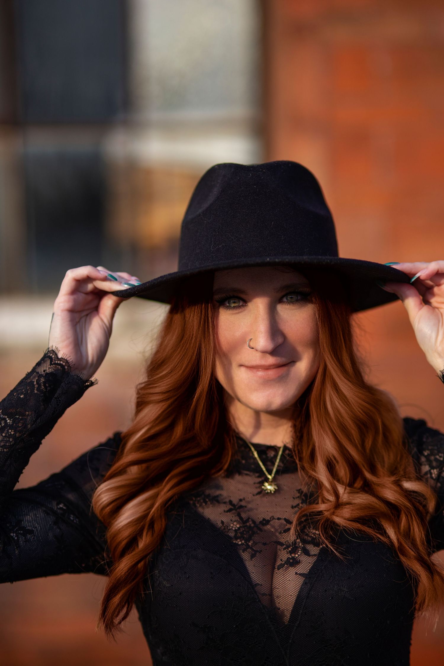 A woman with long red hair wearing a black wide-brim hat and lace dress poses outdoors.