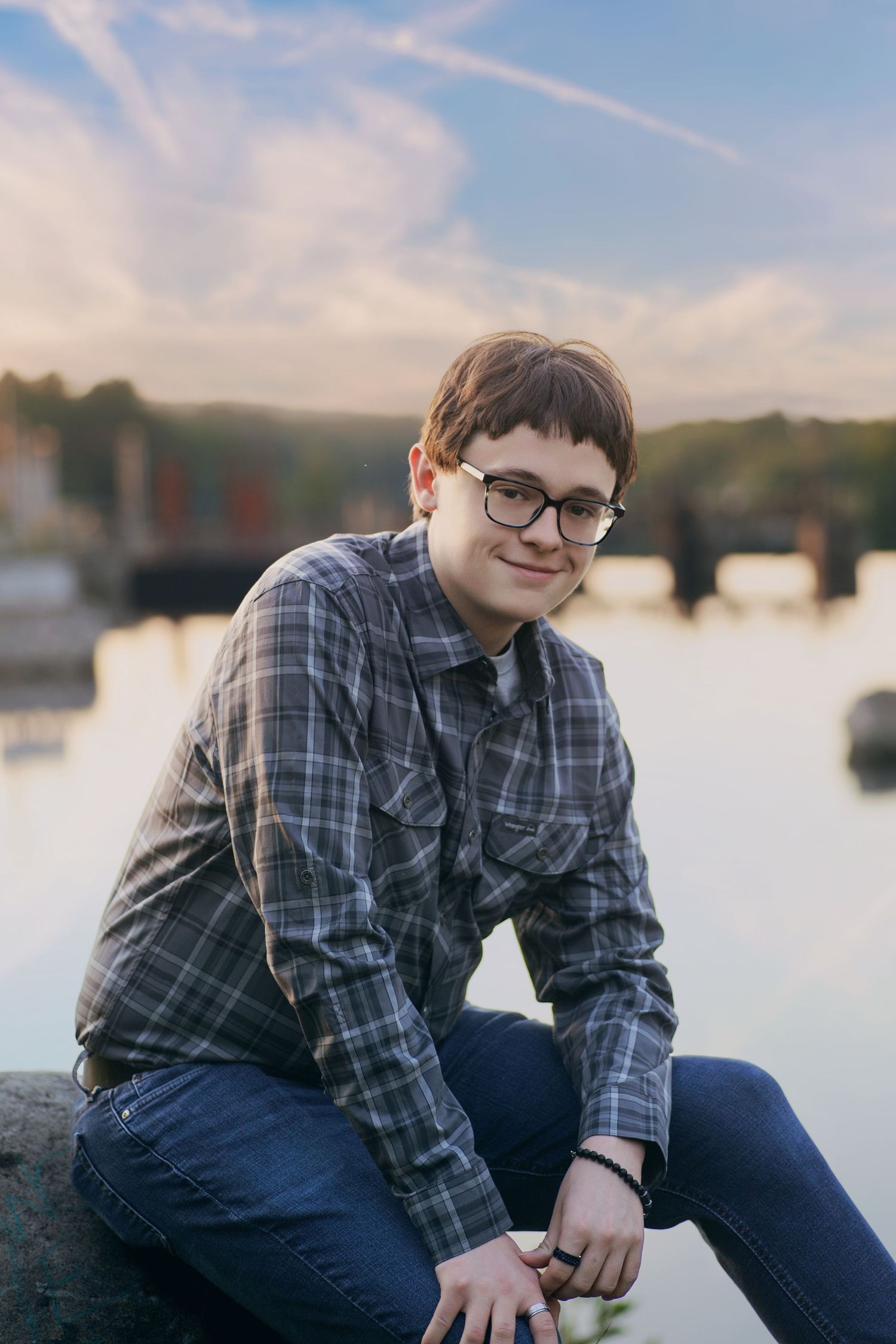 Young musician in plaid shirt and glasses sitting outdoors at sunset by a lake.
