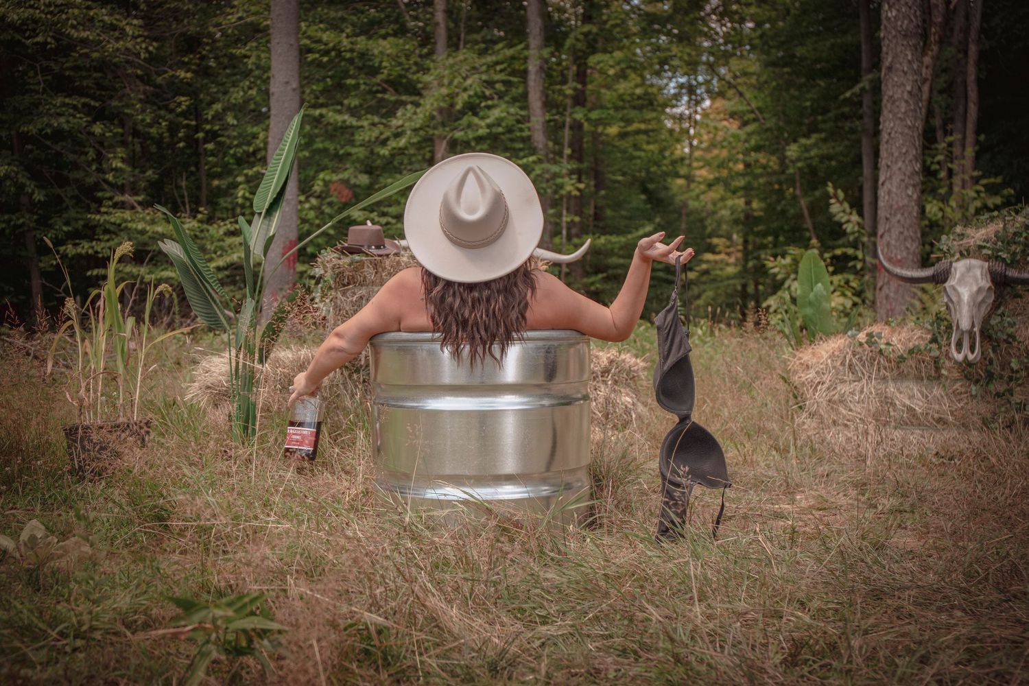A person wearing a cowboy hat sits in a vintage bathtub outdoors in a rustic forest setting with tall trees.