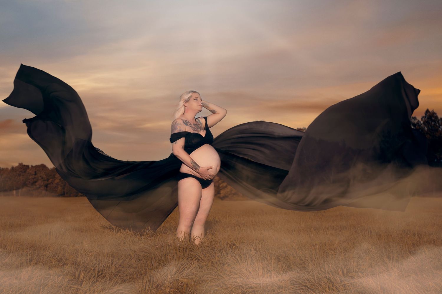 A dramatic maternity photo with flowing black fabric against a sunset sky.