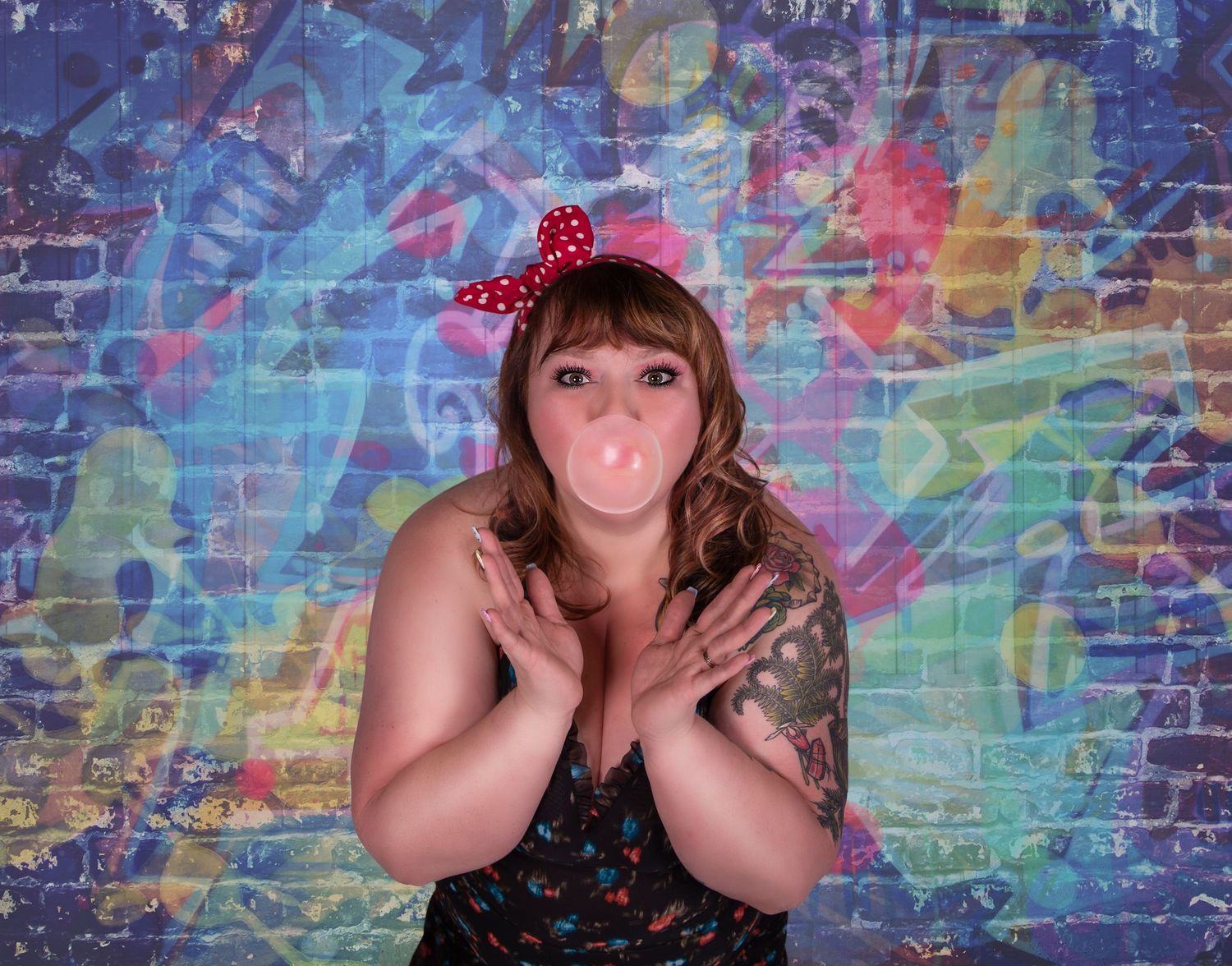 A person wearing a red polka dot bow and floral dress poses playfully against a colorful graffiti wall background.