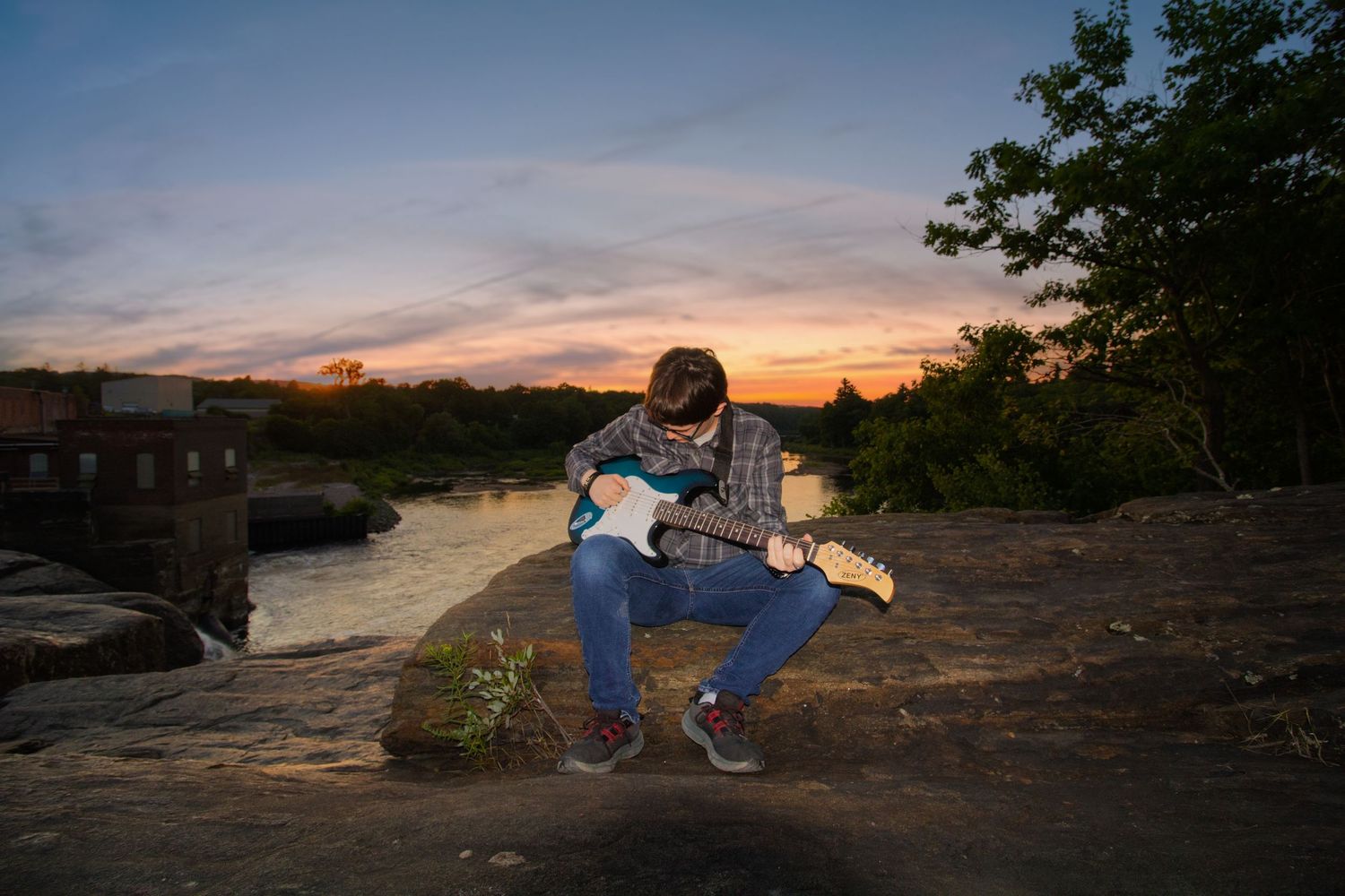 Young man playing electric guitar on rocks by a river at sunset with trees and buildings in the background.