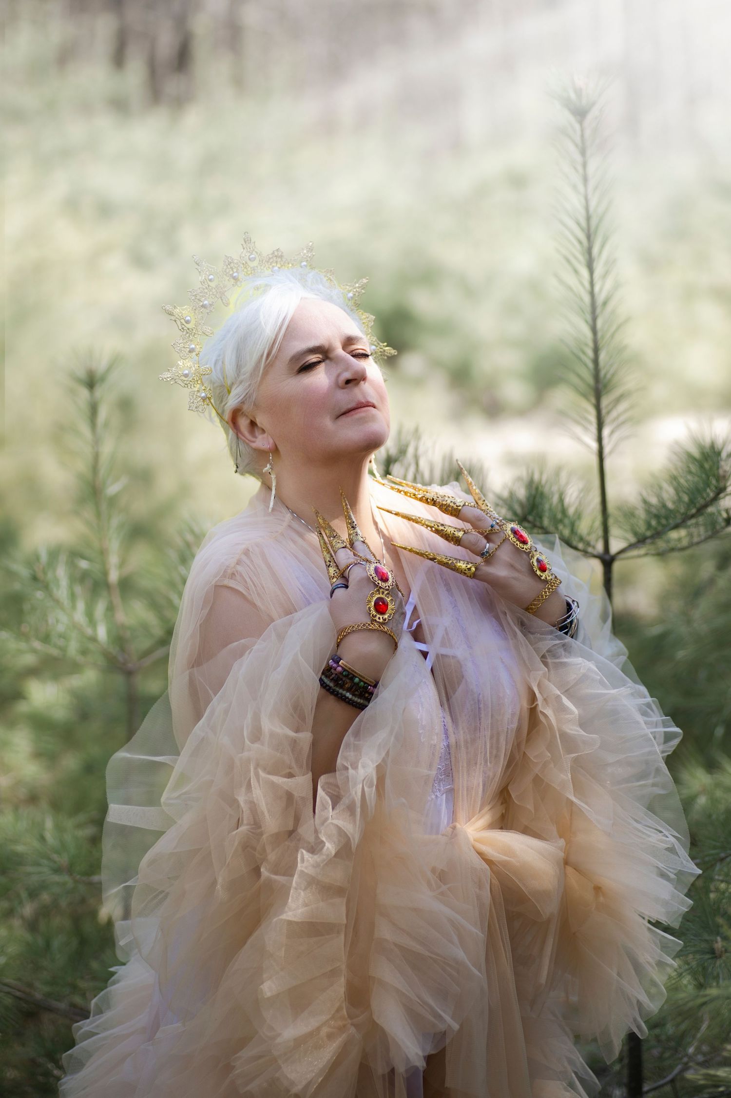 Elegant woman in sheer beige tulle gown with gold jewelry and long nails poses among pine trees in dreamy outdoor setting.