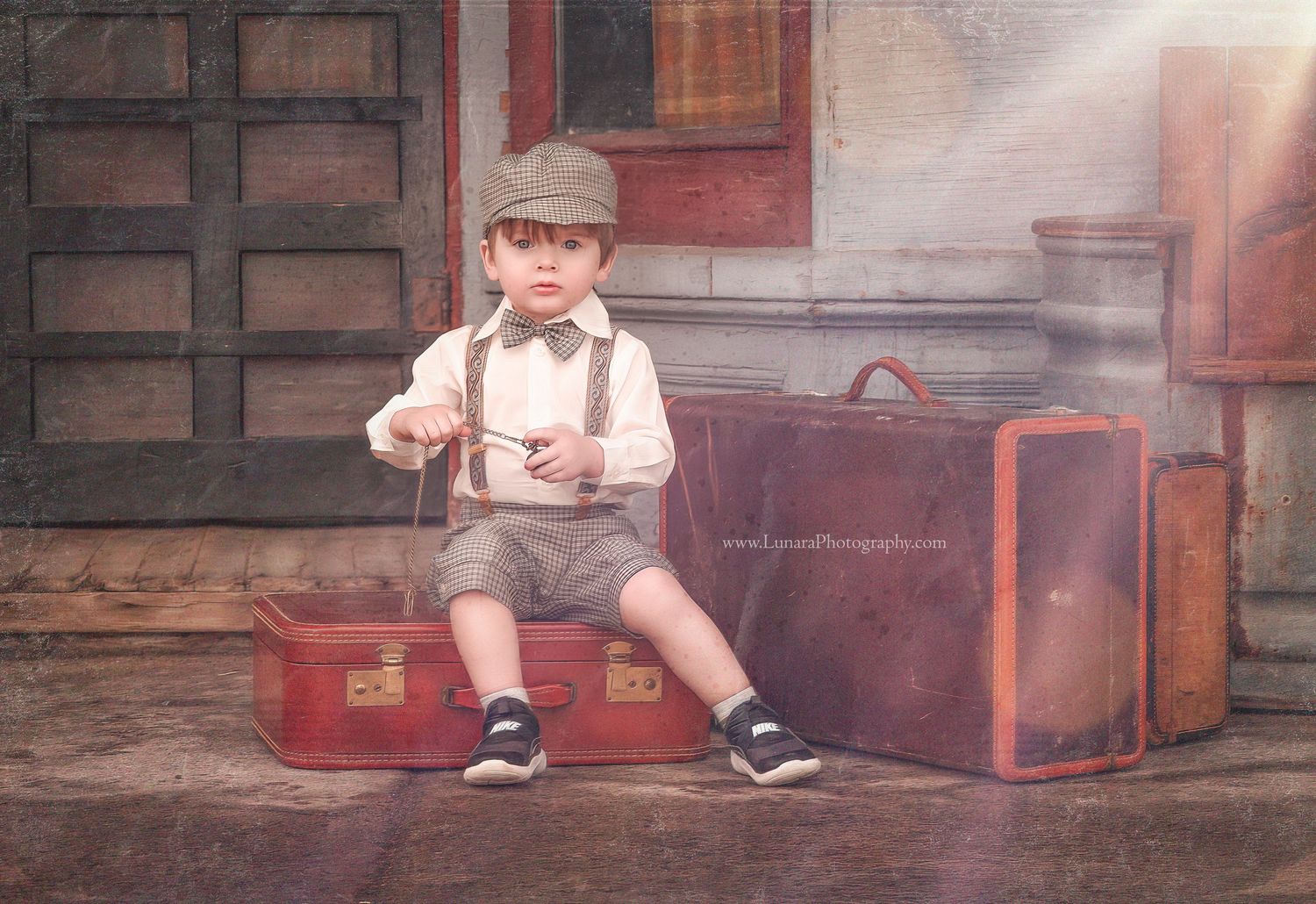 A young boy in suspenders and a newsboy cap sits on vintage red suitcases holding string in a nostalgic portrait.