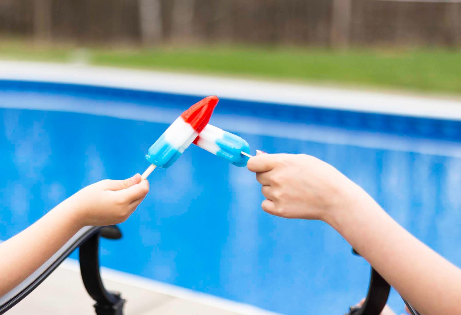 Two hands exchanging a red and white rocket-shaped popsicle in front of a bright blue swimming pool backdrop.