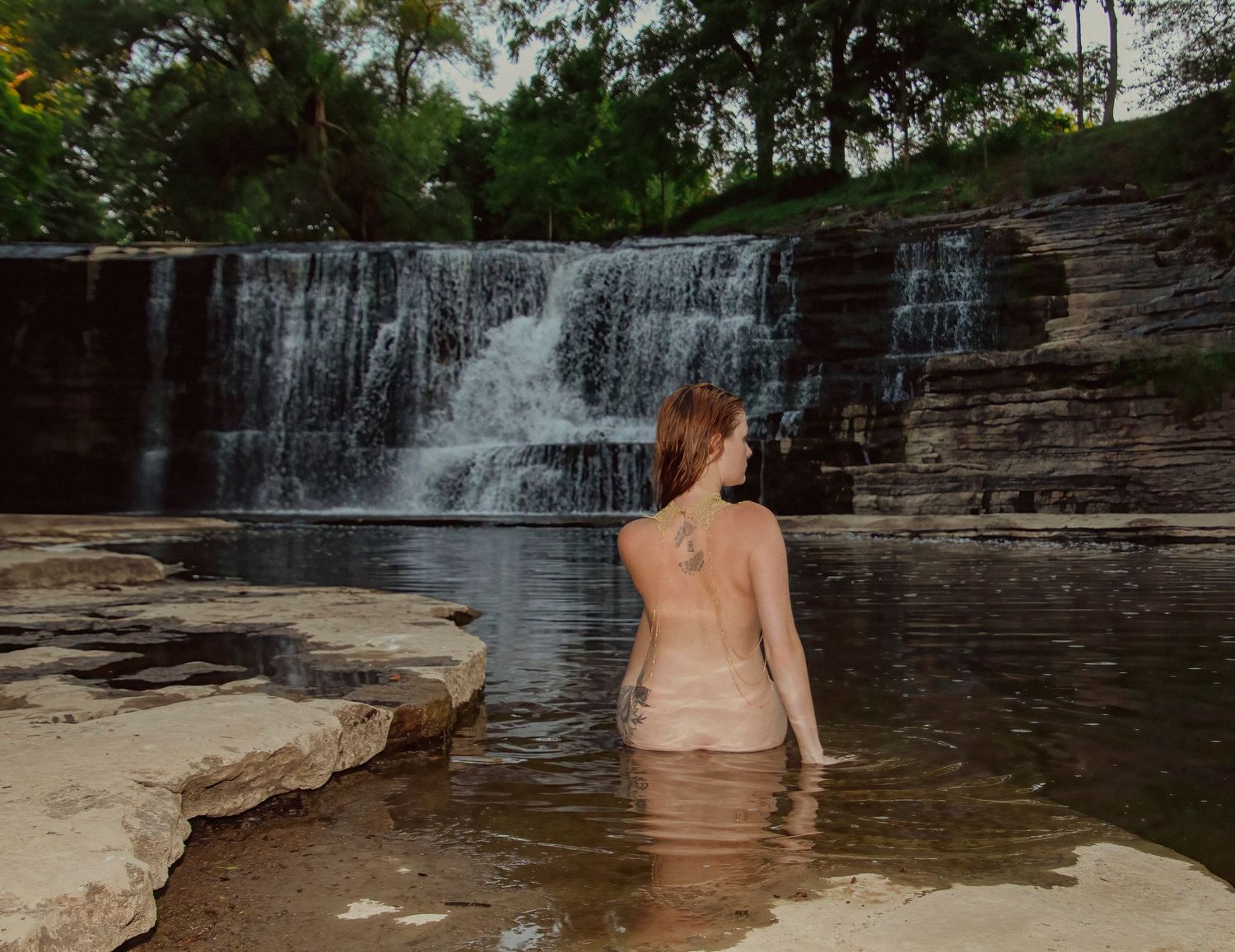 A serene waterfall cascades over rocky ledges into a shallow pool surrounded by natural stone formations.