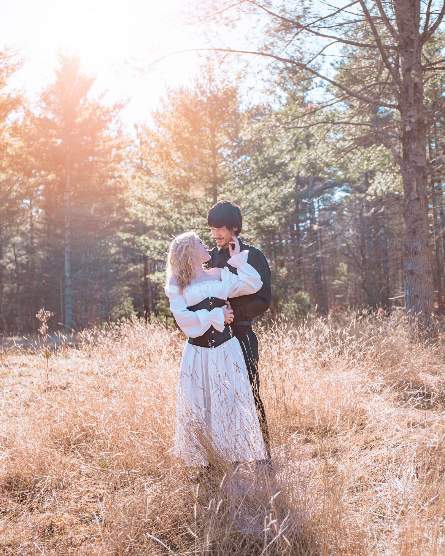 A couple embraces in a romantic moment among tall grass and pine trees at sunset with warm golden light filtering through.