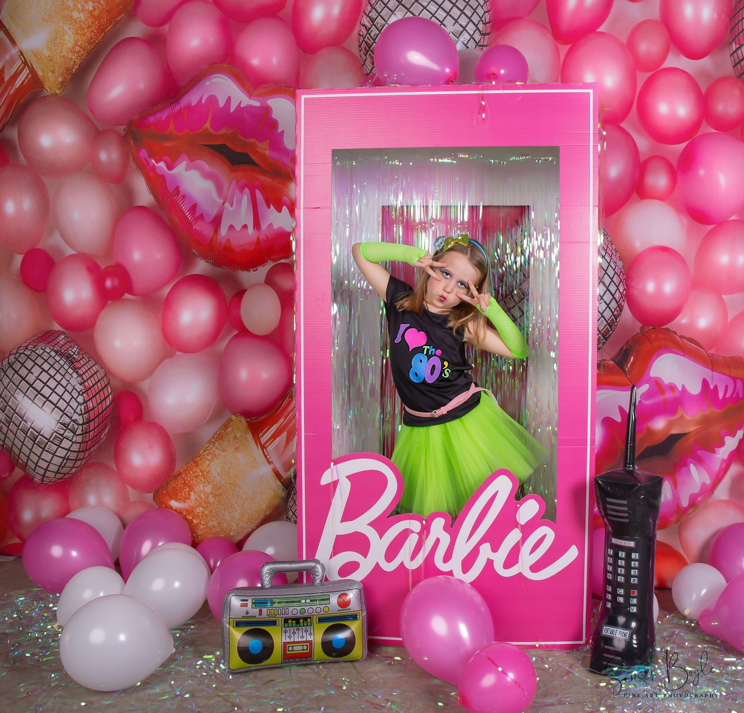 Girl poses in Barbie box prop surrounded by pink balloons, disco ball, boombox, and colorful Barbie-themed party decorations.