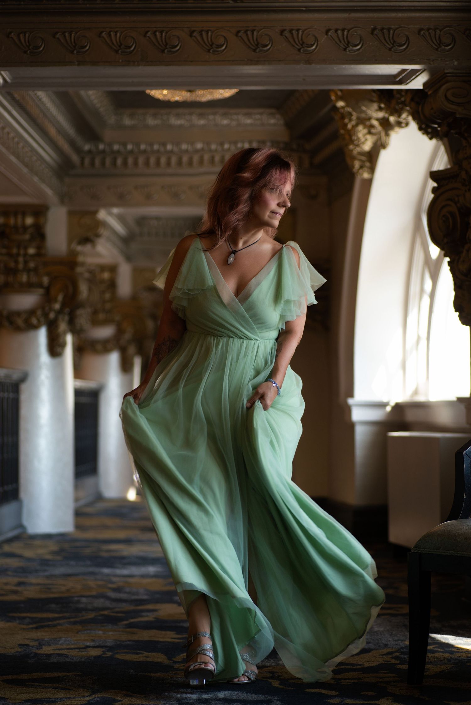 Woman in flowing mint green dress twirls gracefully in a grand historic room with ornate columns.