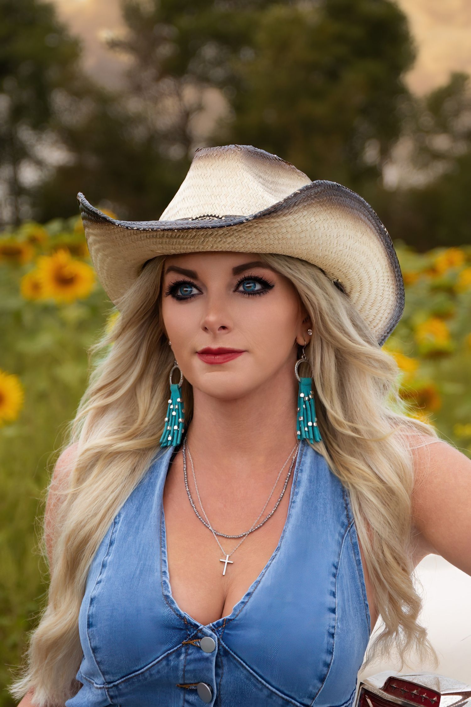 A person wearing a cowboy hat, turquoise earrings and denim top poses in a sunflower field on a sunny day.