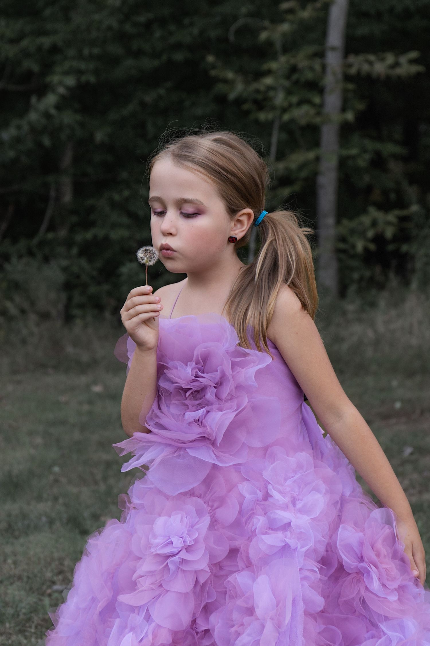 A young person in a flowing purple tulle dress twirls playfully outdoors against a dark forest backdrop.