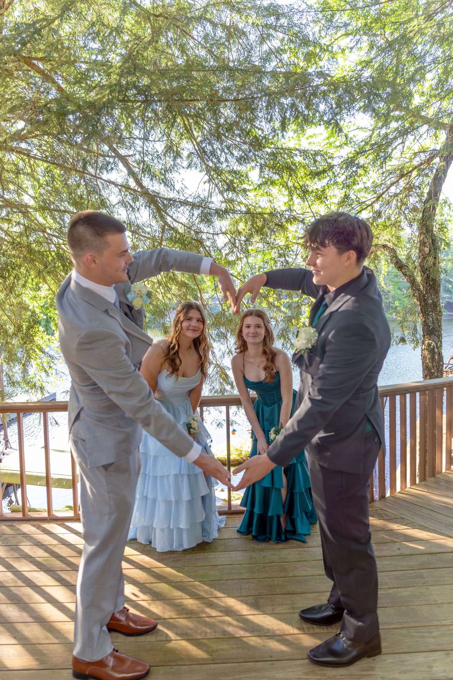 Two grooms hold hands on a wooden deck while guests in formal attire watch during an outdoor wedding ceremony.