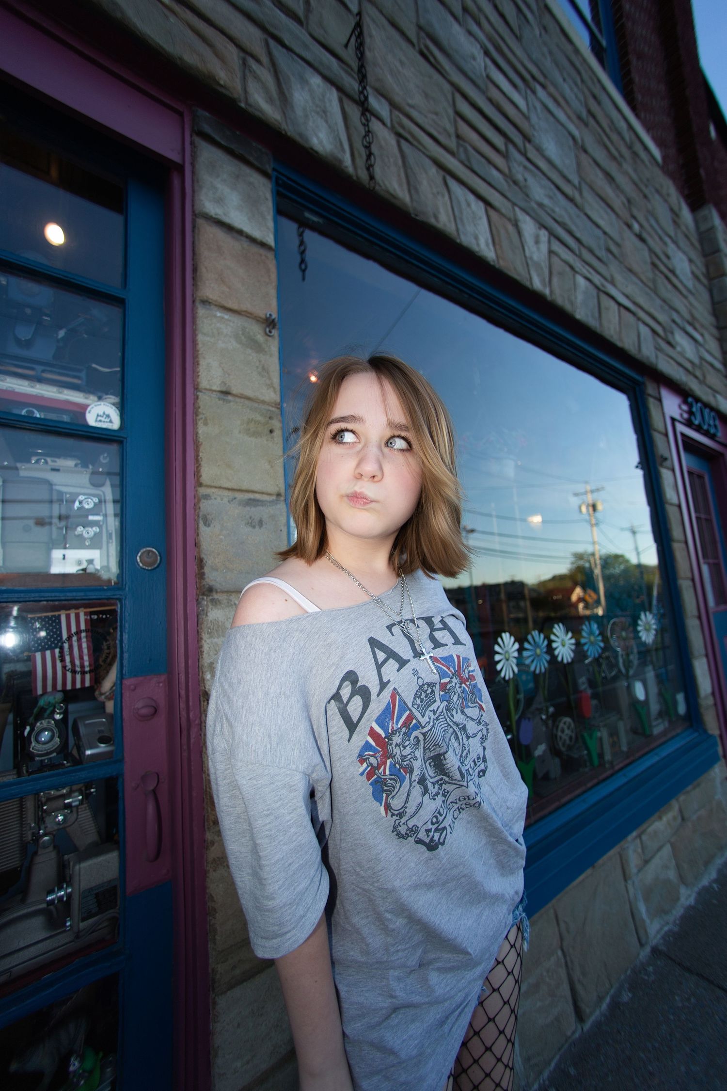 Young woman in gray BATIK graphic tee poses outside a brick storefront with blue trim windows.