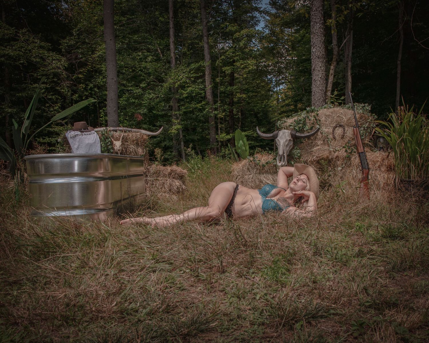 A rustic outdoor scene with hay bales and metal barrels in a wooded forest setting at dusk.