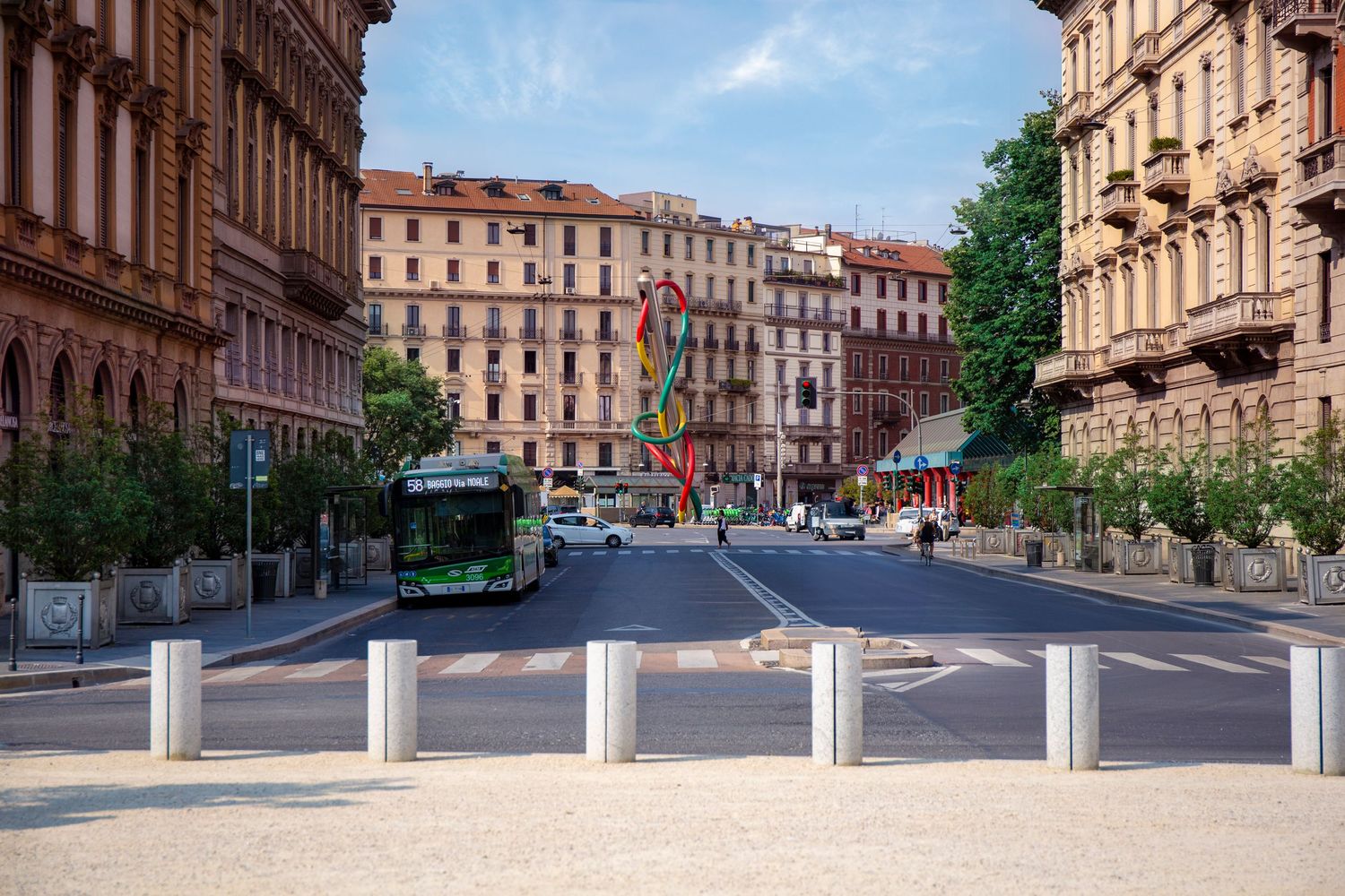 A green bus drives down a European city street lined with historic apartment buildings and retractable security bollards.