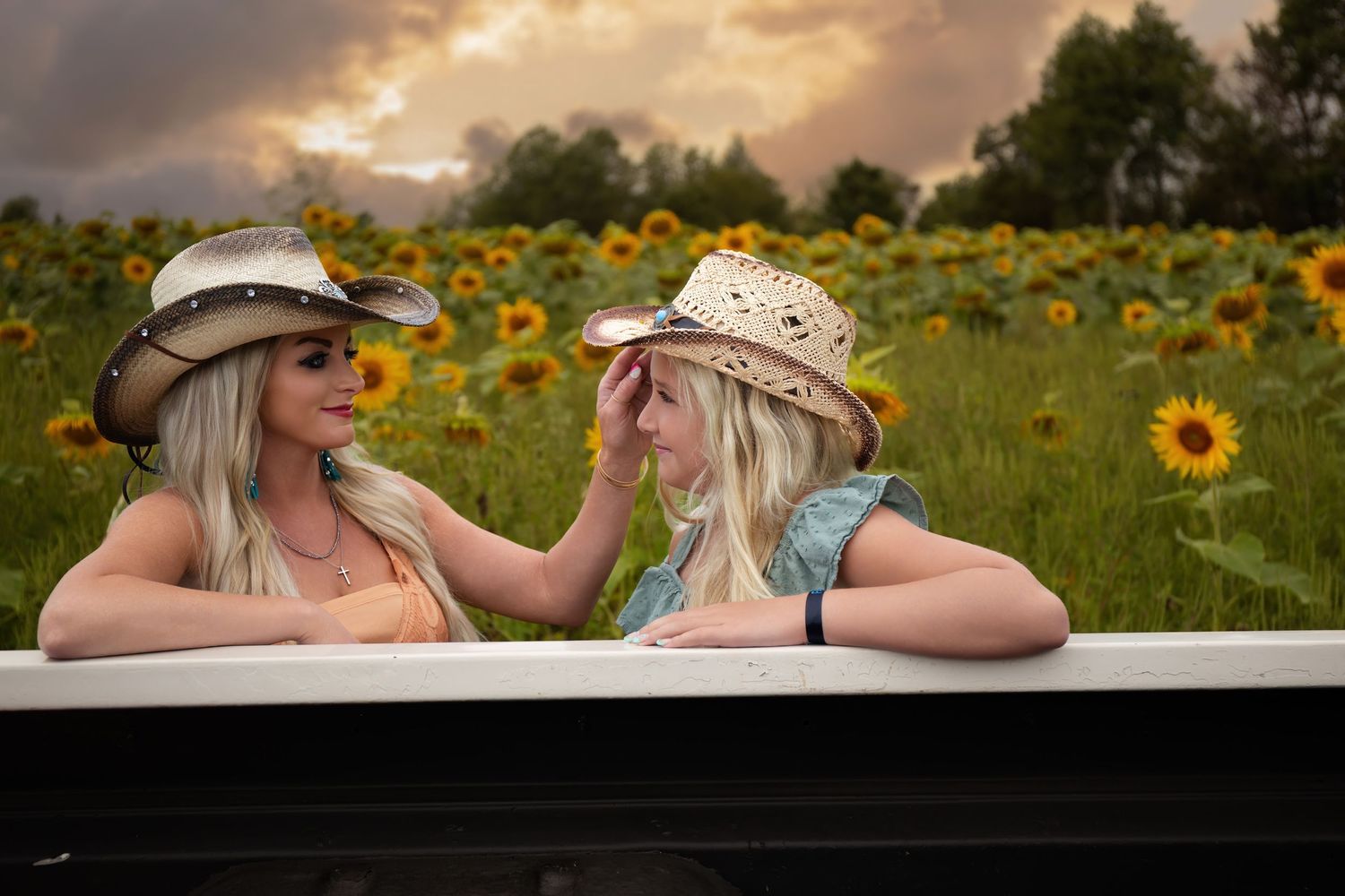 Two women in straw hats smile and lean on white railing in front of vibrant sunflower field at sunset.