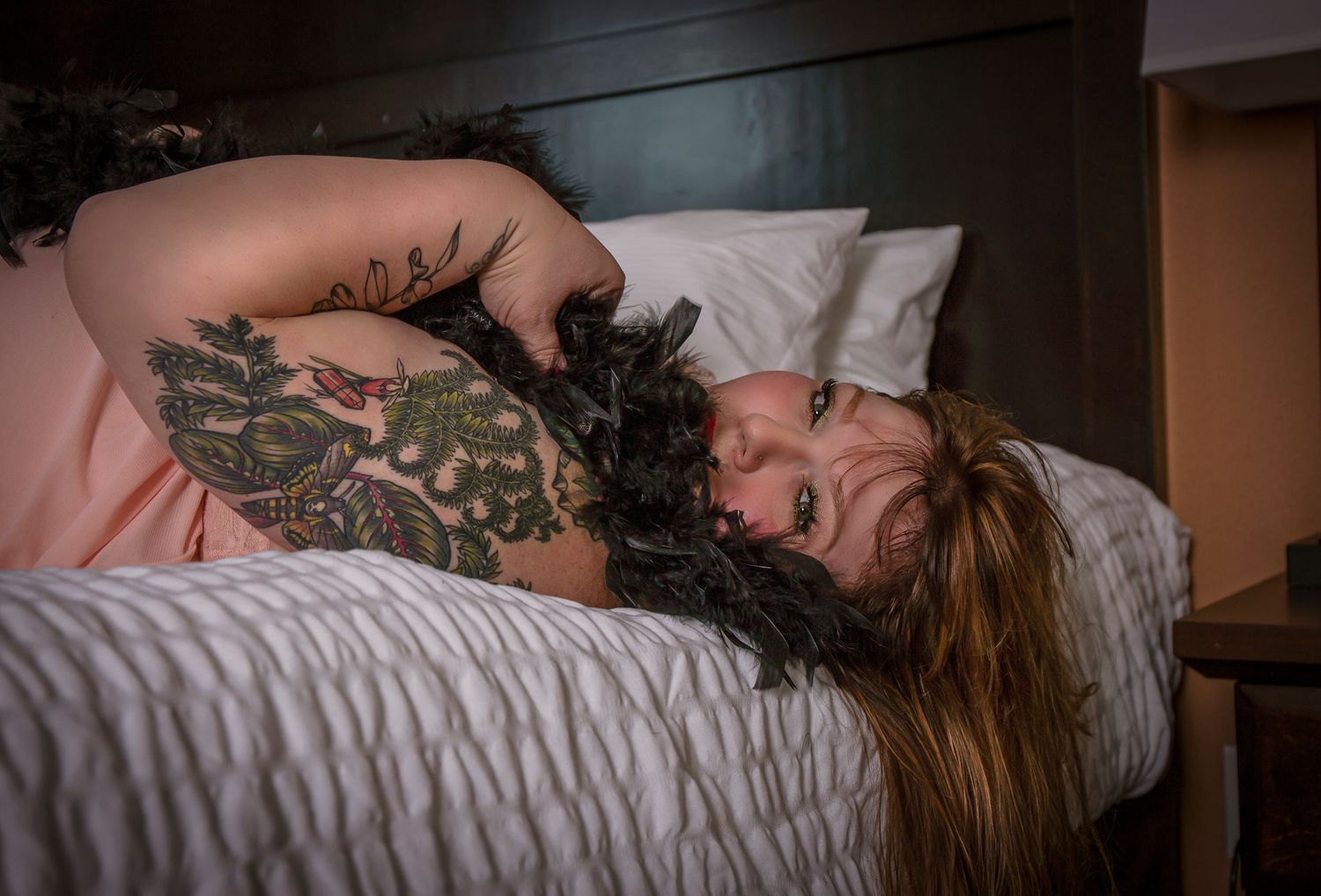 A tattooed person with long hair lays curled up on white bedding in dim lighting.