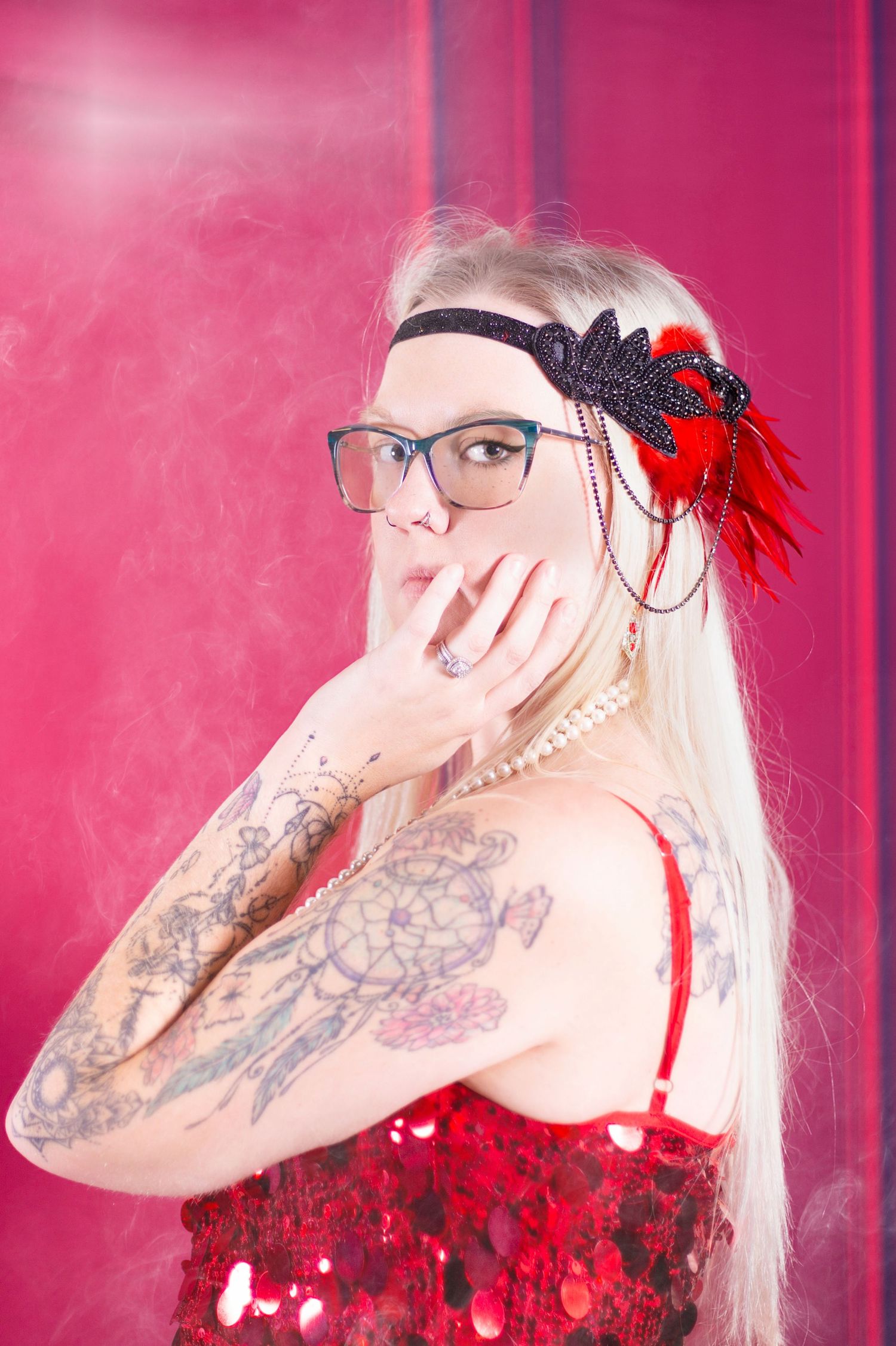 A person with long platinum hair and glasses poses in red lighting wearing a red accessory against a pink background.