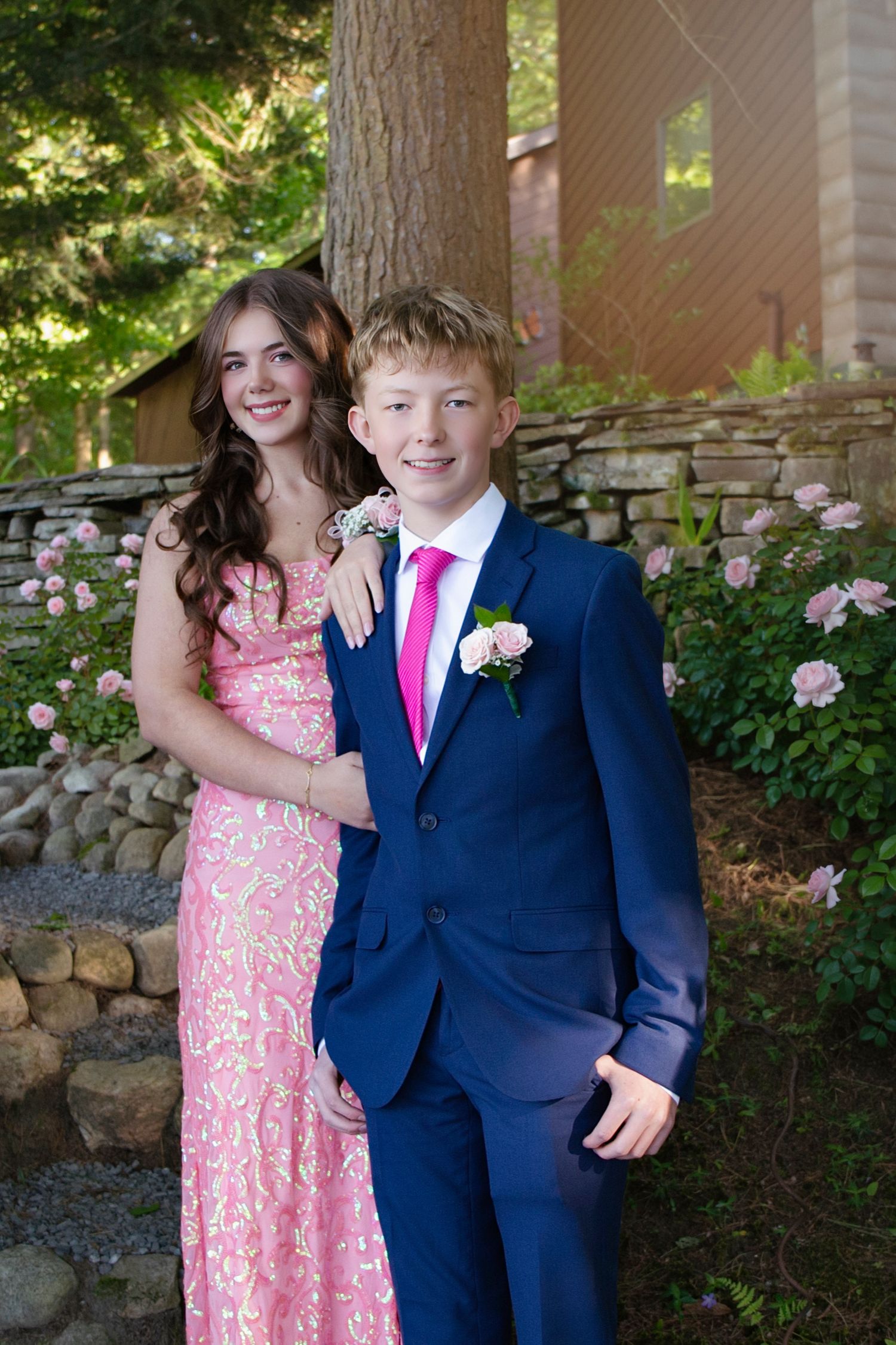 Teen couple dressed for prom in pink floral gown and navy suit with pink tie posing outdoors.