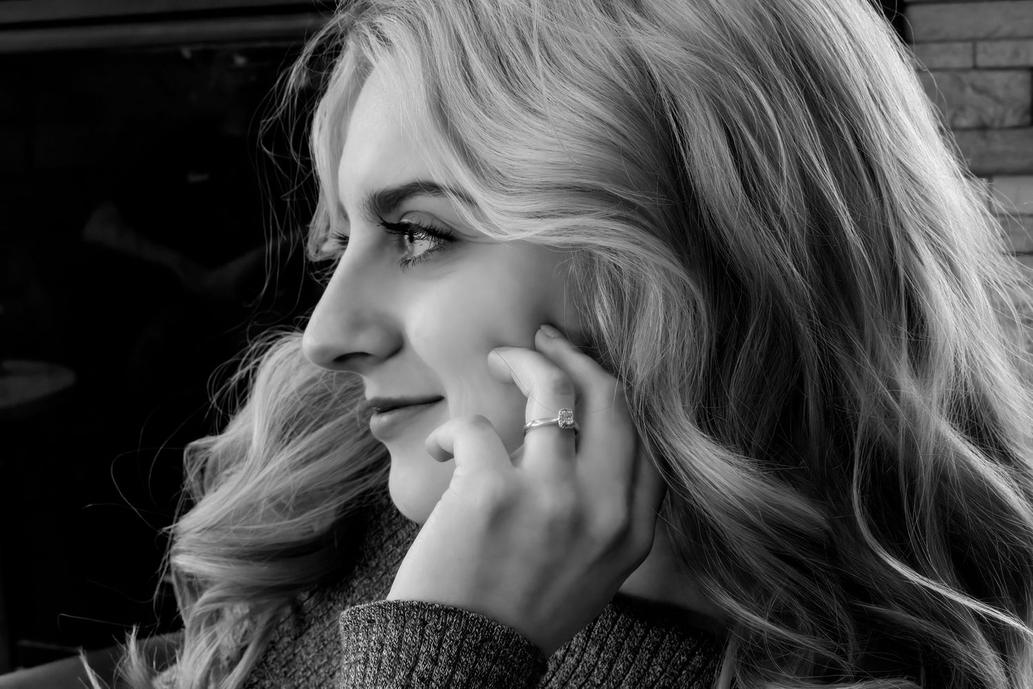 Black and white portrait of a blonde woman touching her face, wearing a ring, looking sideways thoughtfully.
