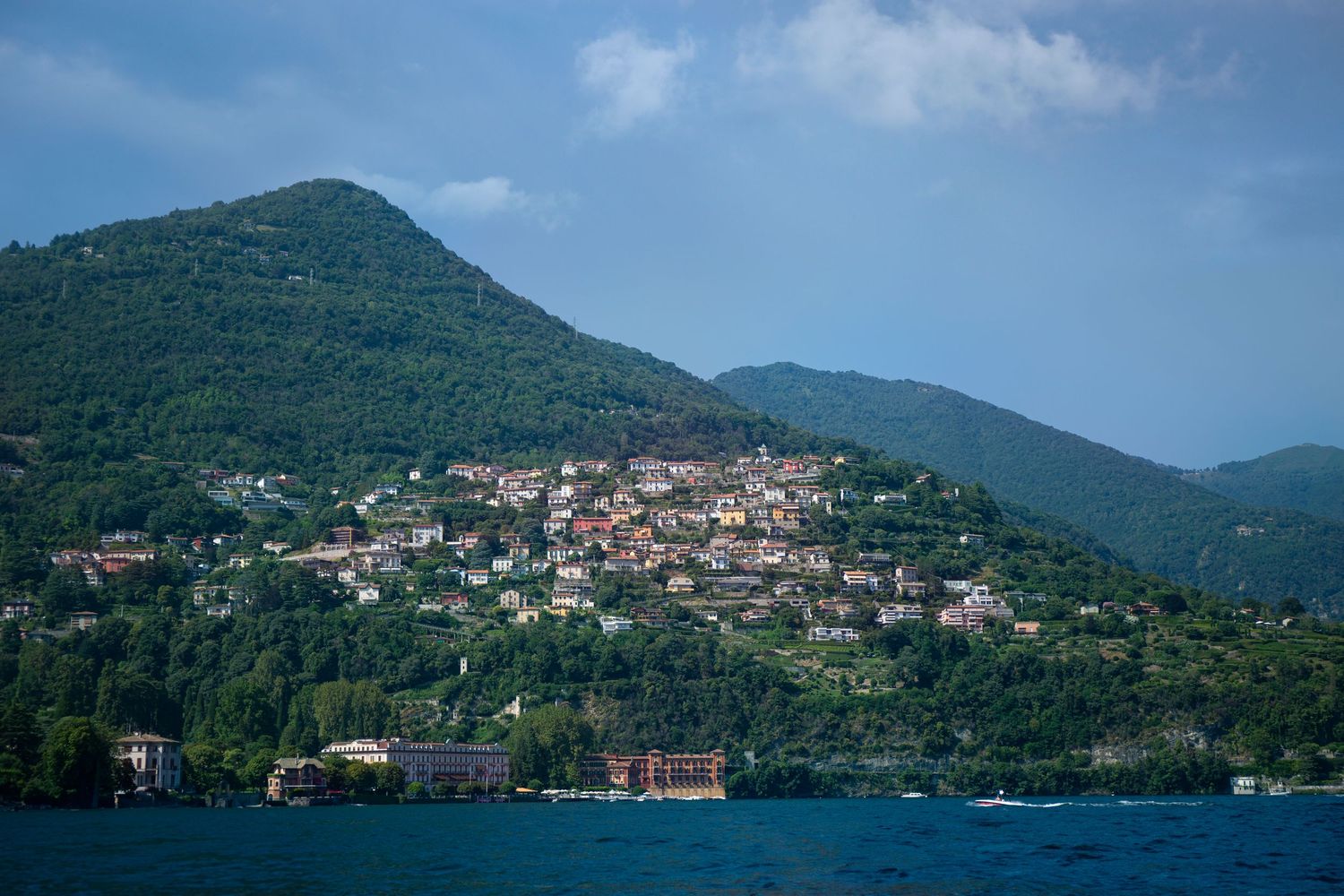 Mediterranean coastal village nestled on green hillside overlooking blue waters with mountains in background.