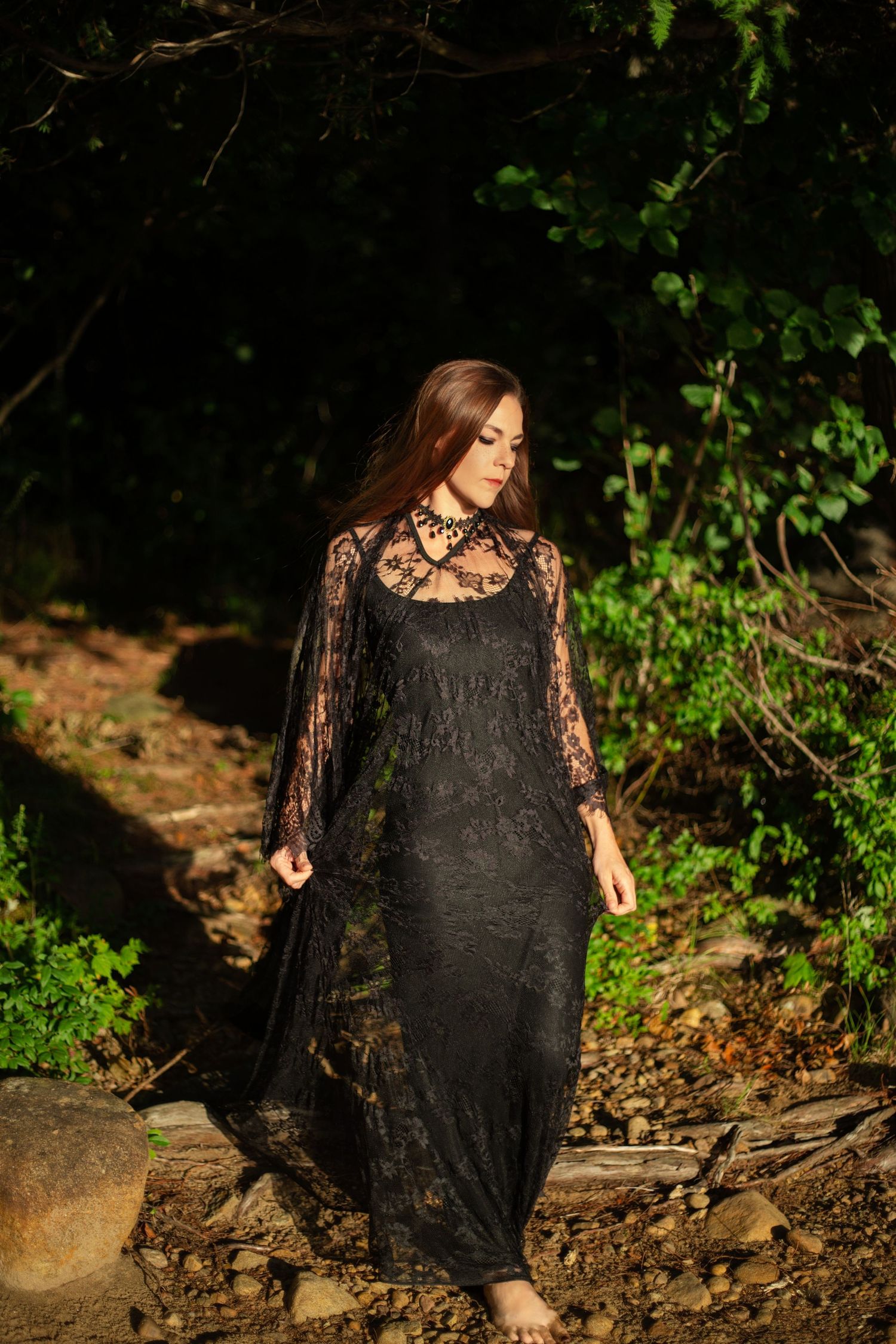 Woman in black lace dress poses dramatically in a wooded outdoor setting.