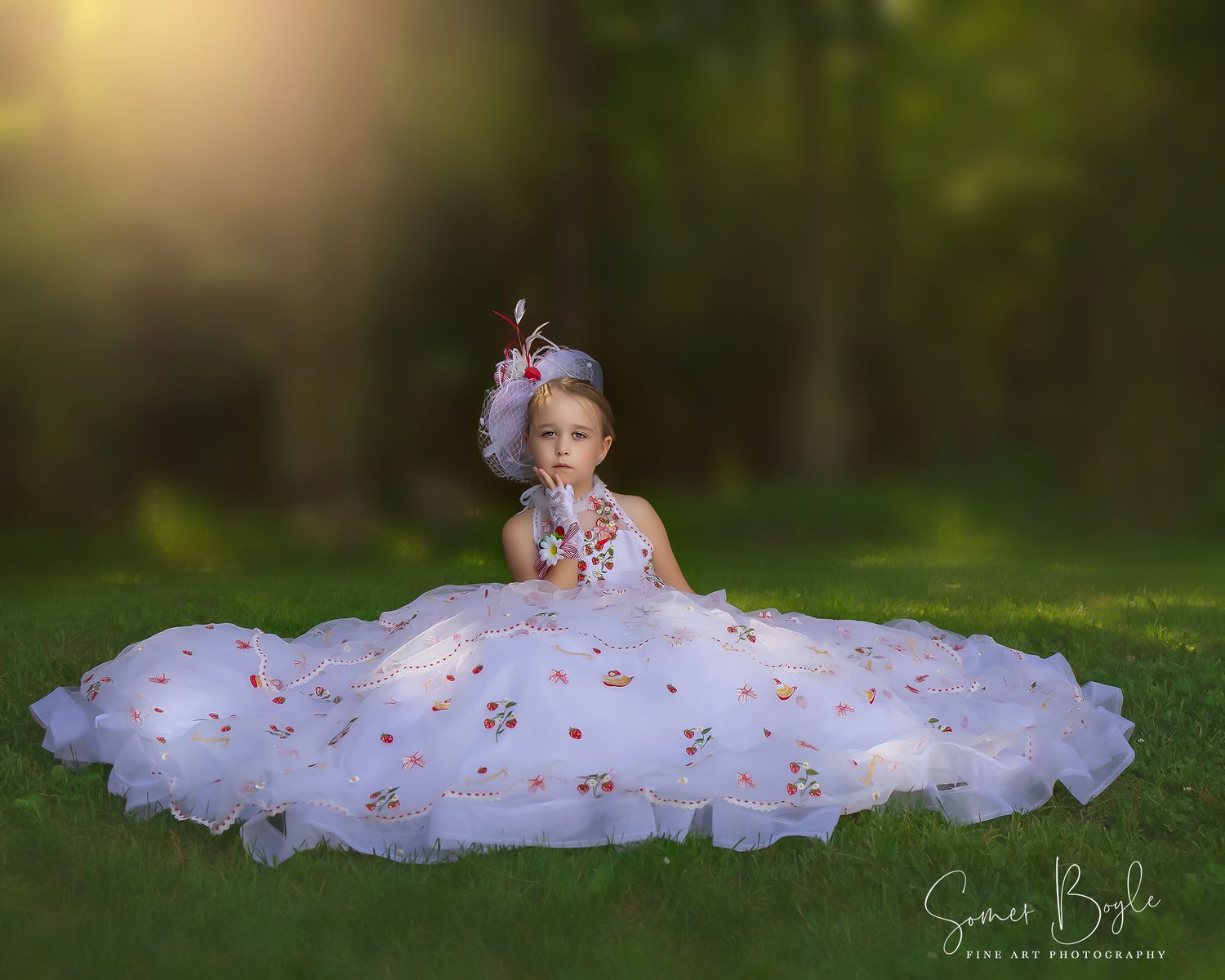 Young girl in white floral ballgown sits on grass in golden sunlight with floral headpiece.