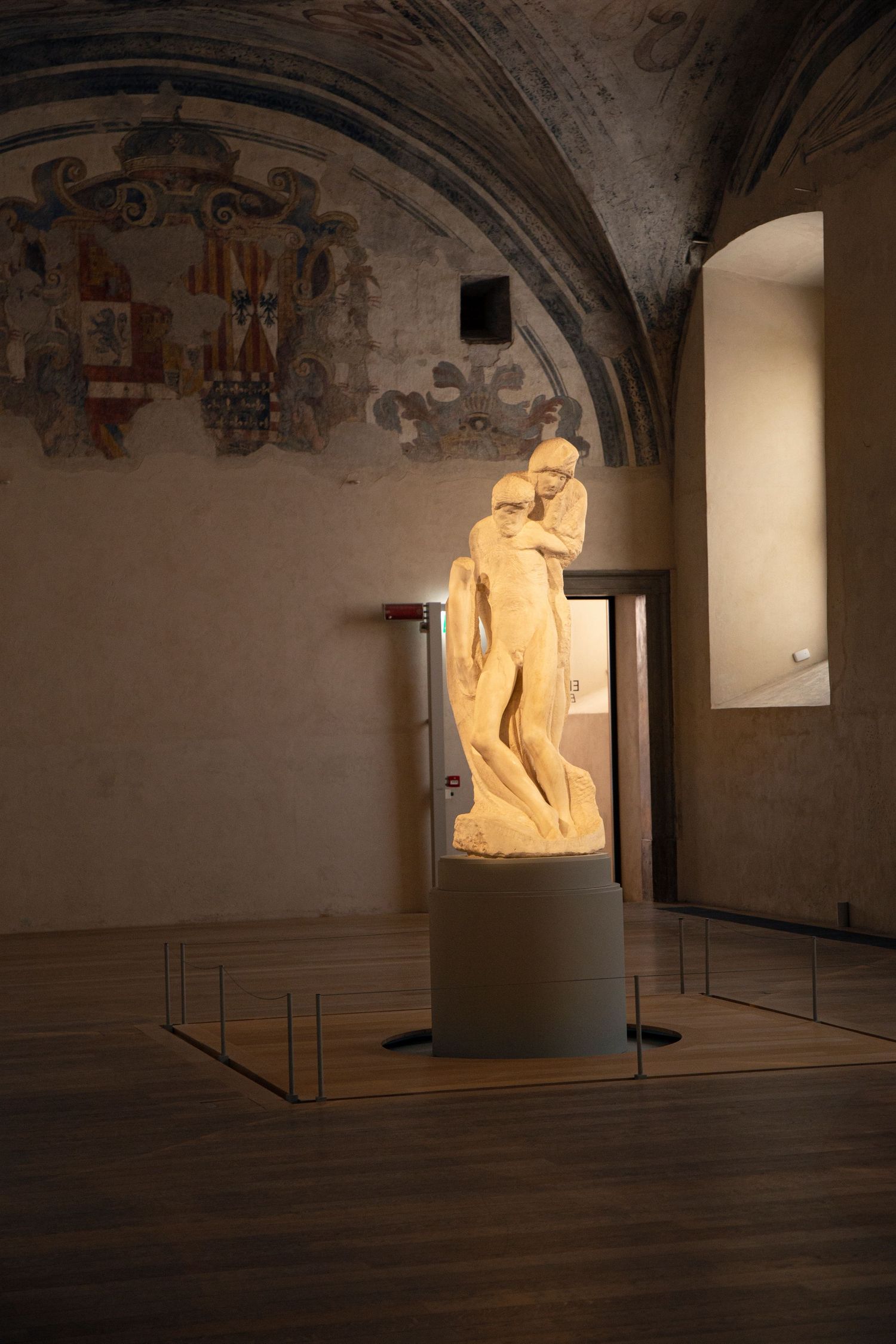 Illuminated marble sculpture displayed in a dimly lit vaulted gallery with historic frescoes on the ceiling.