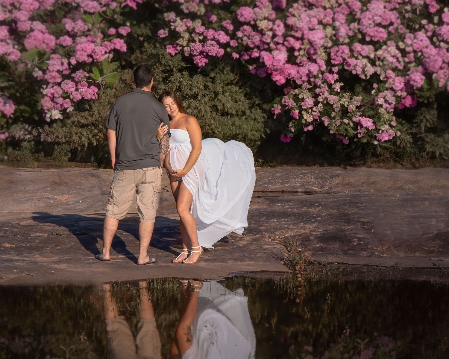 A romantic couple's reflection in water with pink flowering bushes in the background.