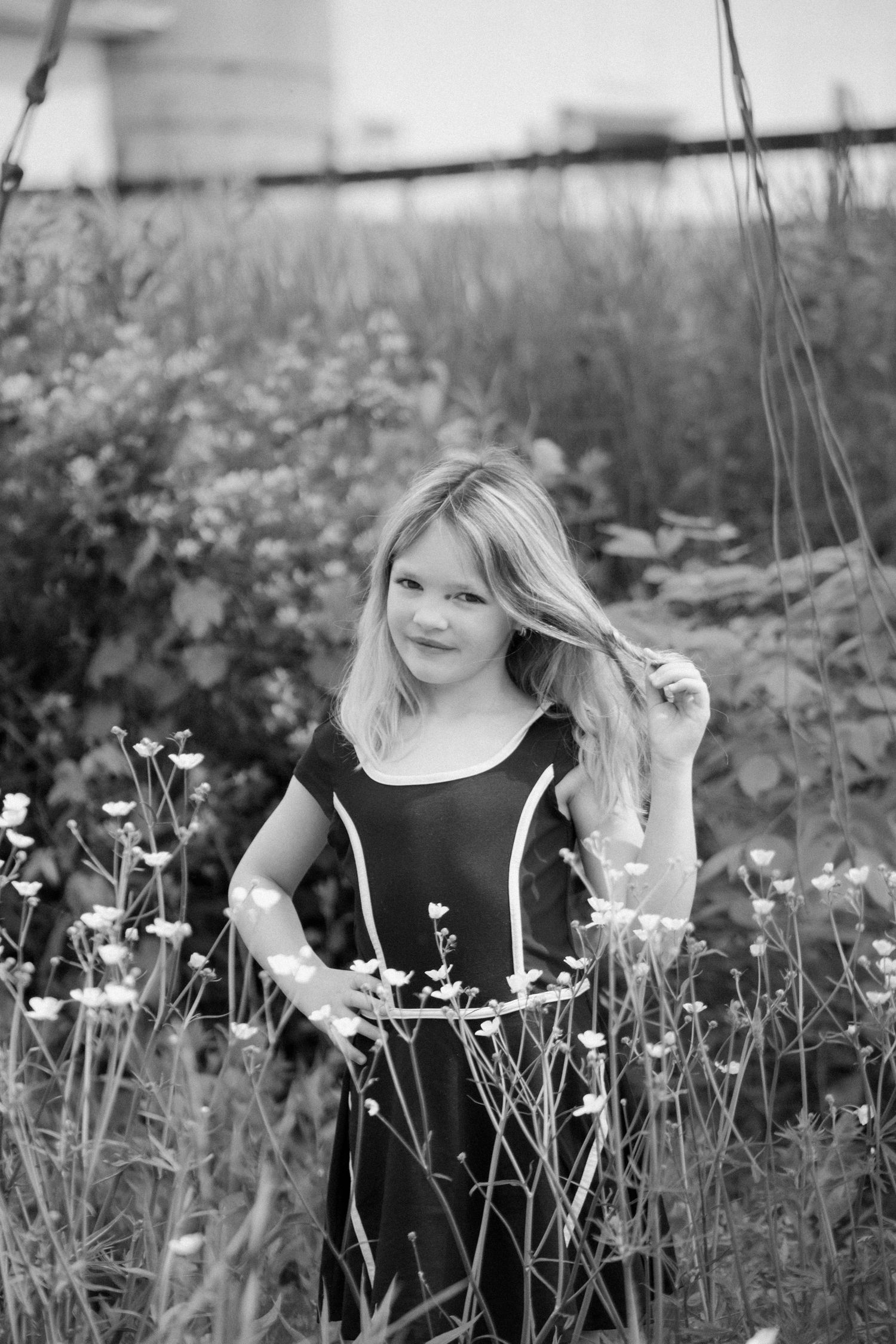 Black and white photo of a young blonde girl in a dark dress standing among wildflowers in a garden.