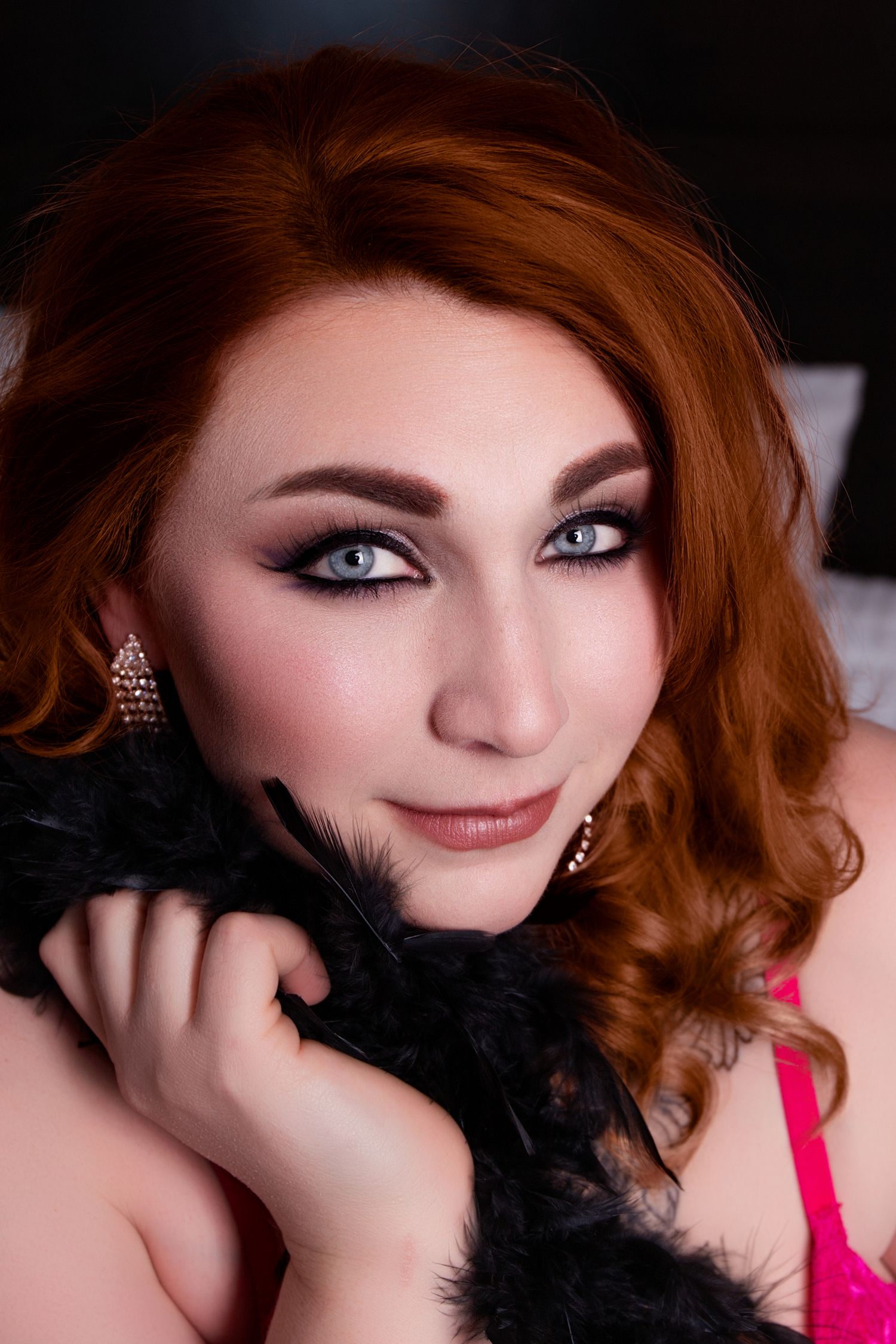 Portrait with vibrant red hair, dramatic eye makeup, and black feather boa against dark background.