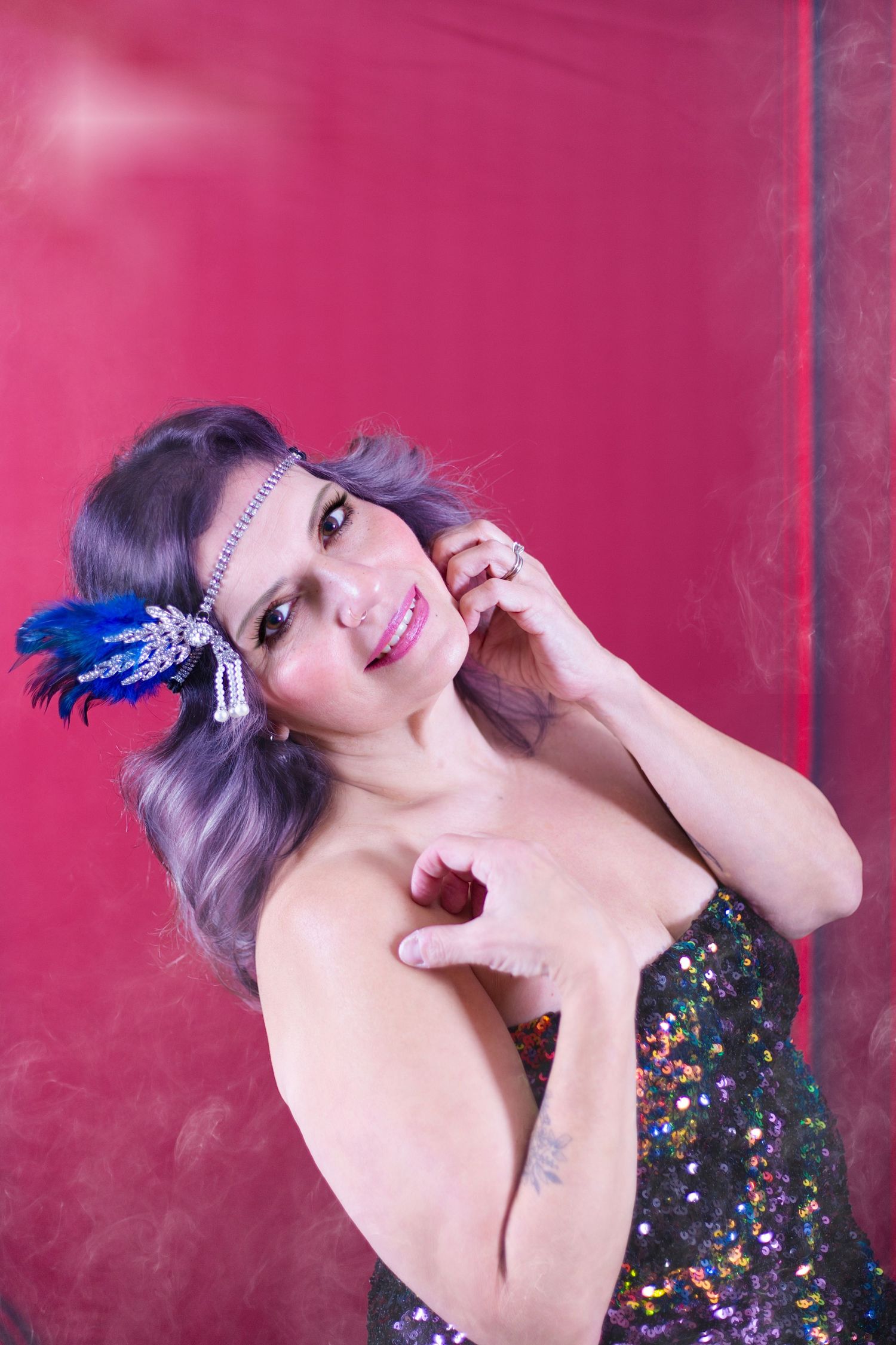 A burlesque performer in a sequined dress and blue feather hair accessory poses against a pink backdrop.