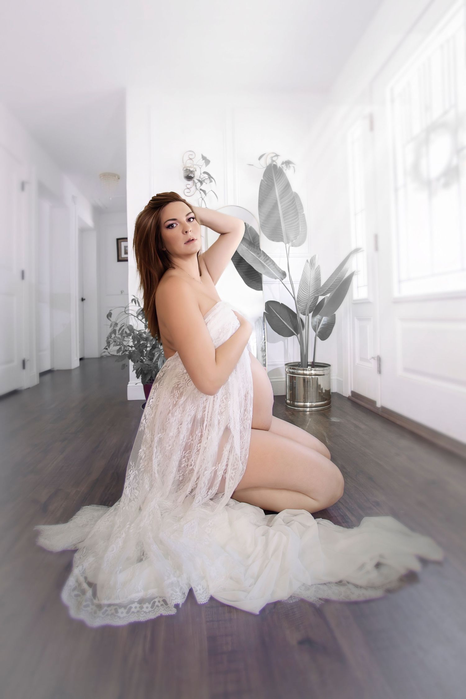 Pregnant woman in white lace robe kneeling gracefully on hardwood floor in bright white room with potted plants.