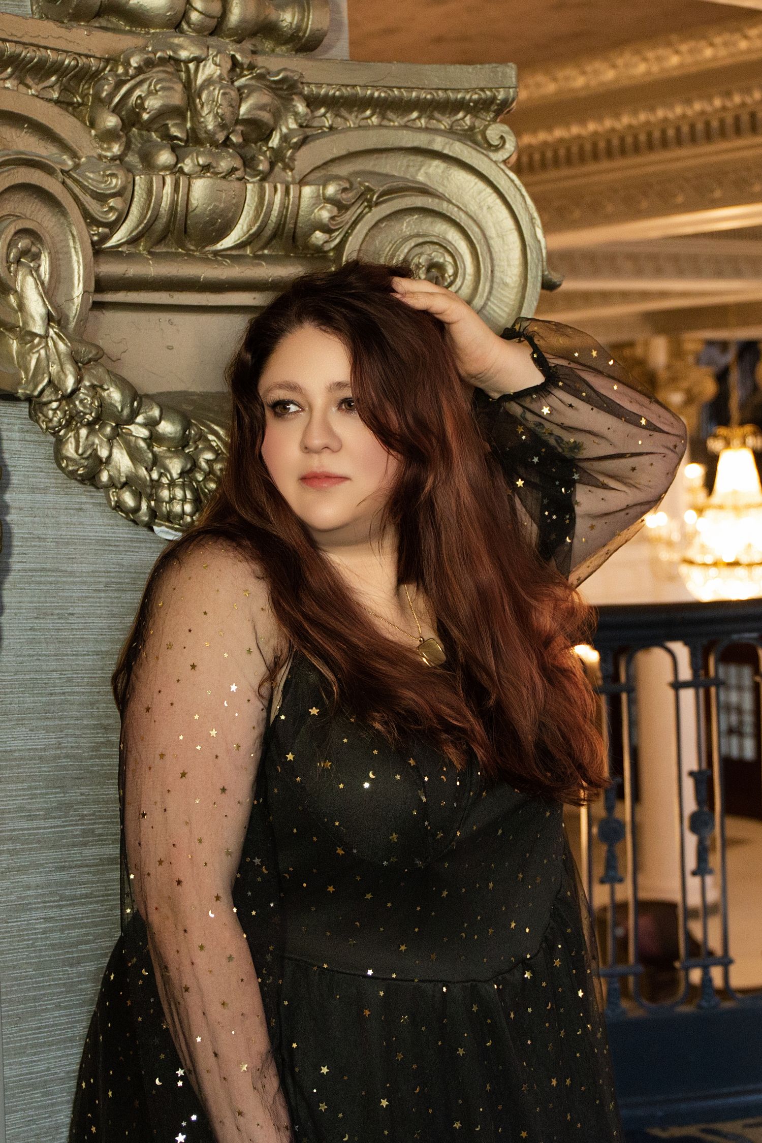 Person in black evening gown with sheer sleeves poses against ornate architectural backdrop.