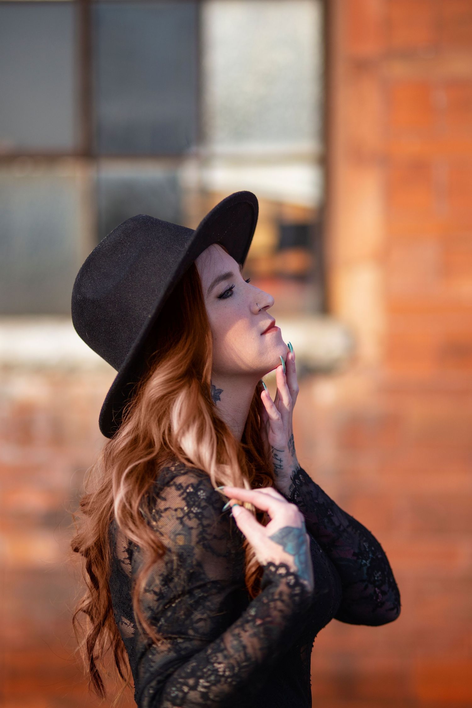 Red-haired woman in black lace dress and wide-brim hat poses outdoors near brick wall.