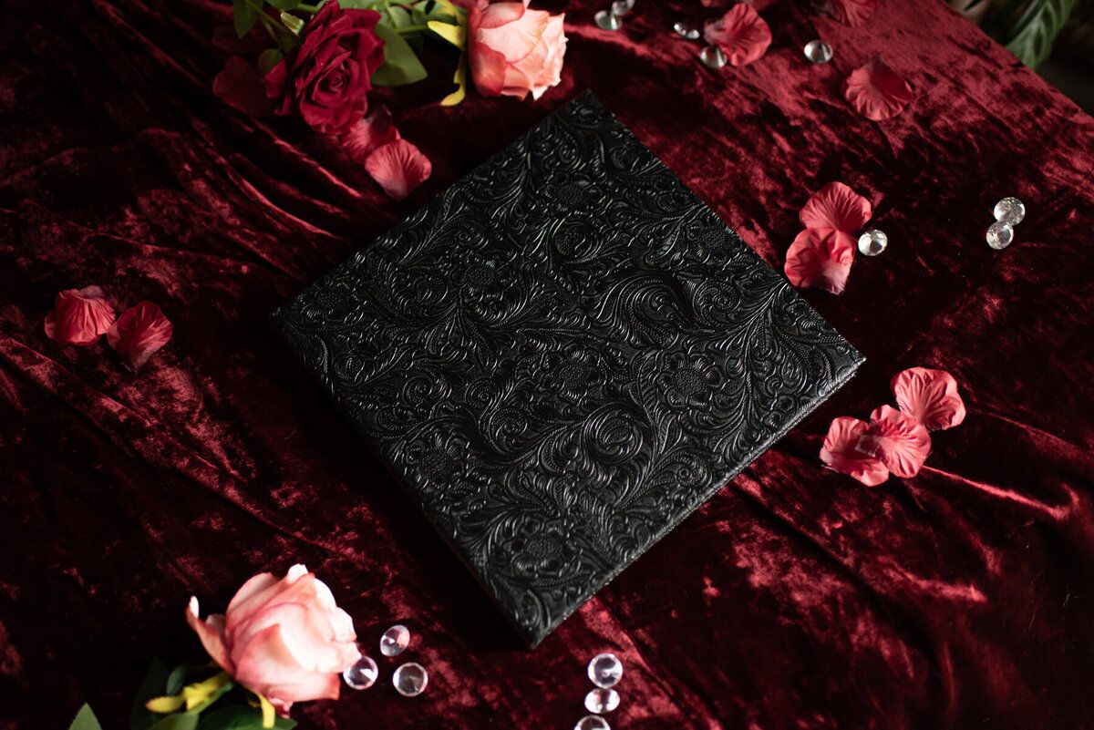 Elegant black photo album with floral patterns displayed with pink roses on dark velvet.