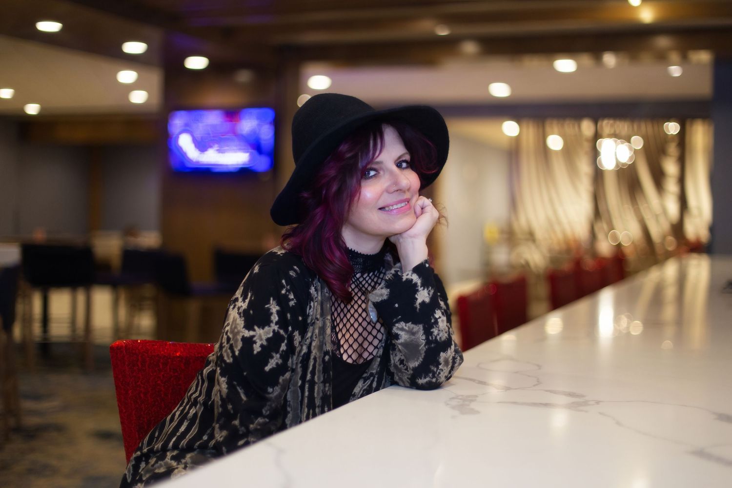 A person with dark hair sits in a dimly lit room with warm lighting and red seating visible in the background.