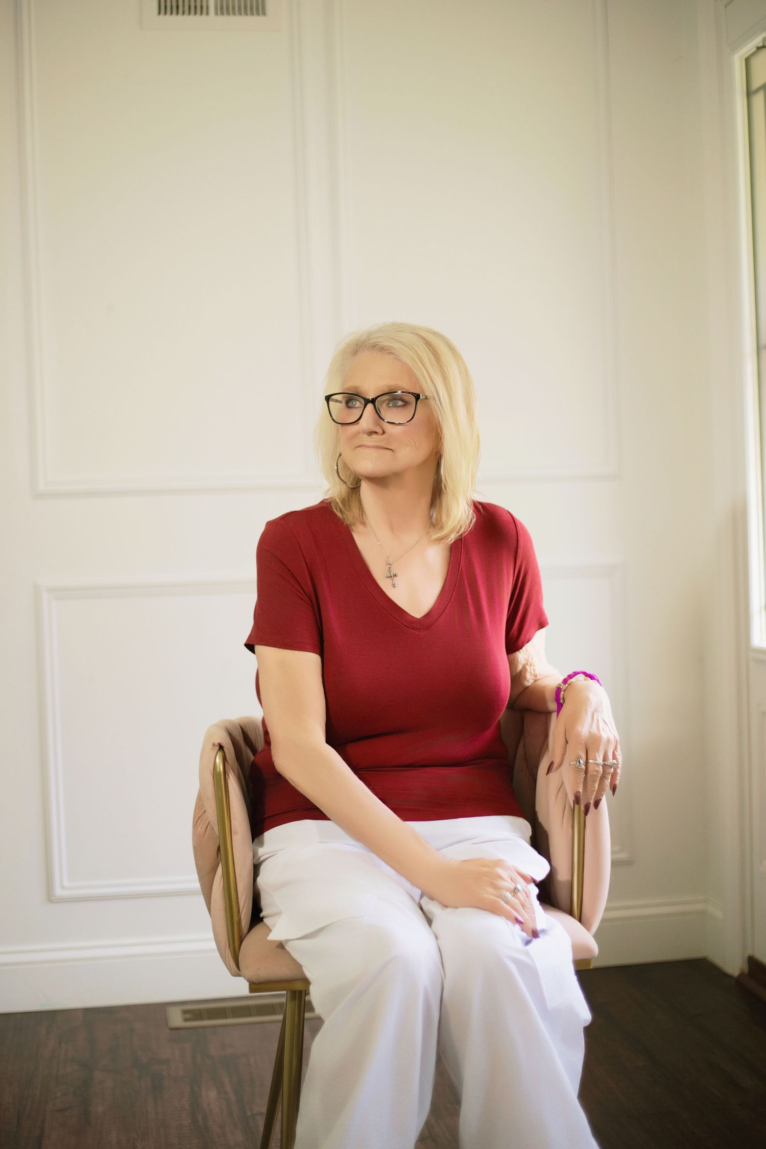 A figure in a red top and white pants sits thoughtfully in a chair against a light colored wall.