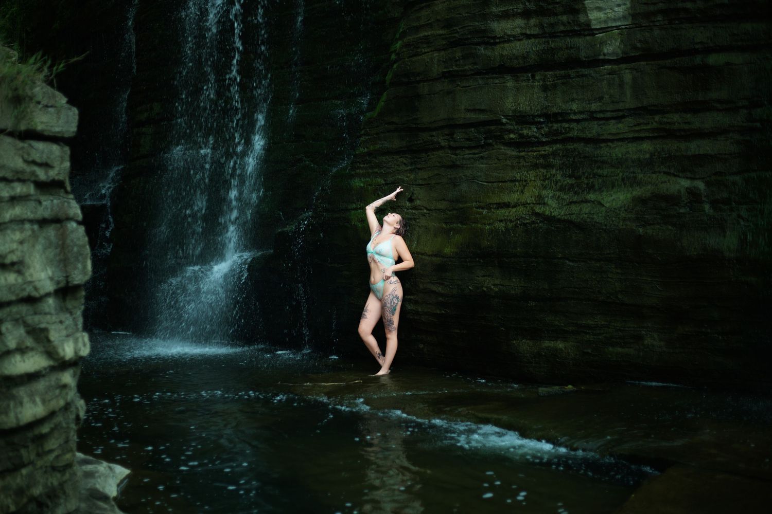 A person stands beneath a cascading waterfall in a dark rocky gorge, creating a dramatic silhouette against the flowing water.
