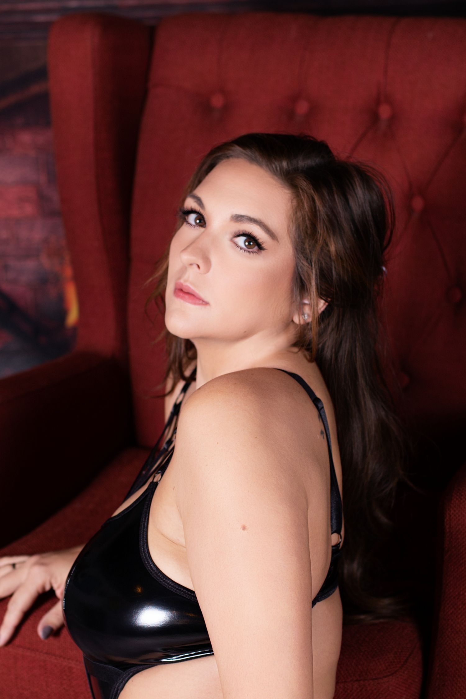 A person in a black latex top poses on a red tufted couch in a dimly lit room.