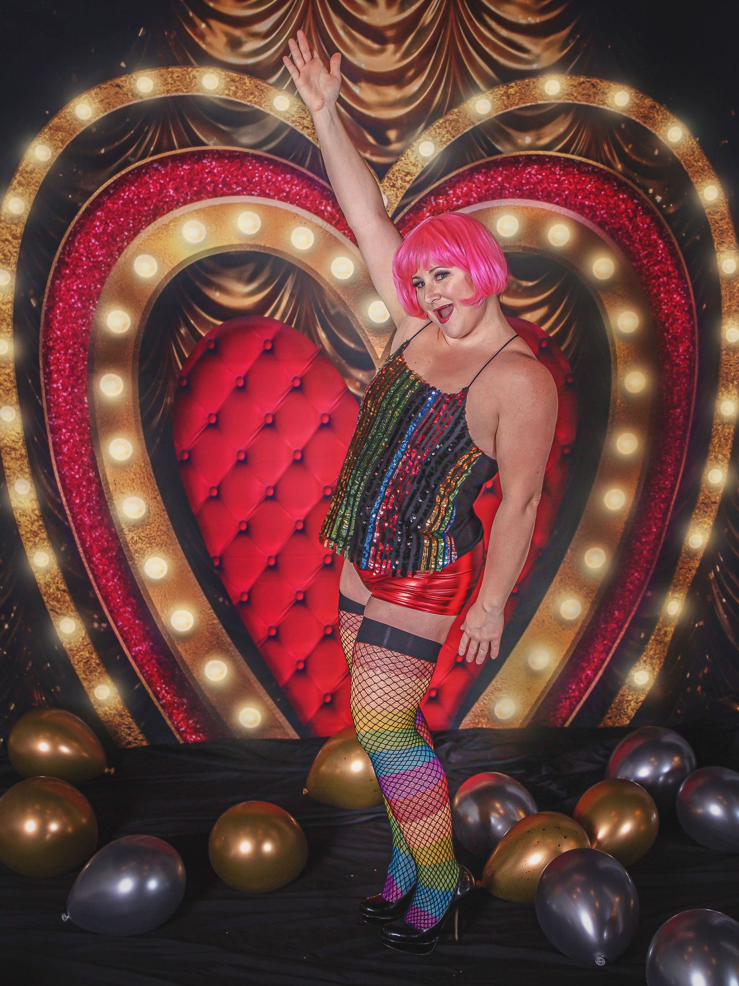 Burlesque performer in rainbow sequin outfit poses in front of illuminated heart-shaped backdrop with red tufted cushion.