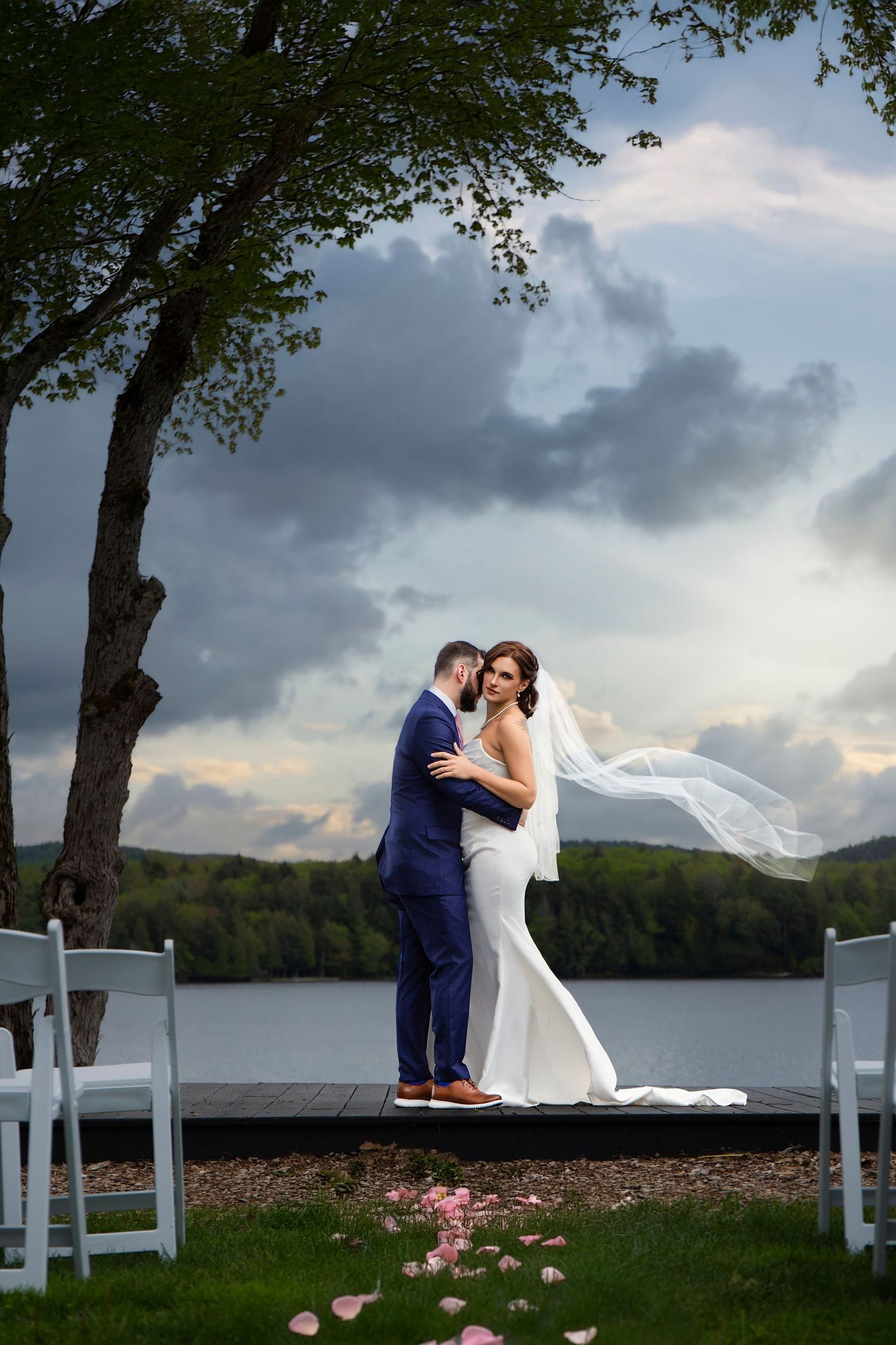 Romantic wedding couple embracing on white chairs by lakeside with dramatic cloudy sky.