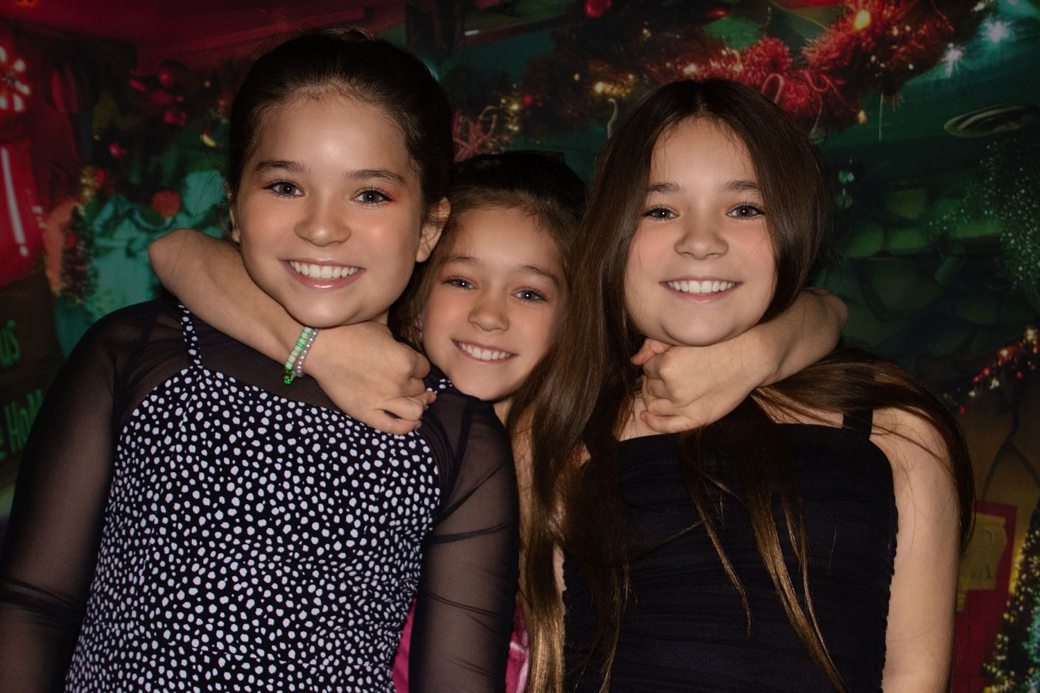 Three people smile together in a dimly lit setting with decorative lighting in the background.