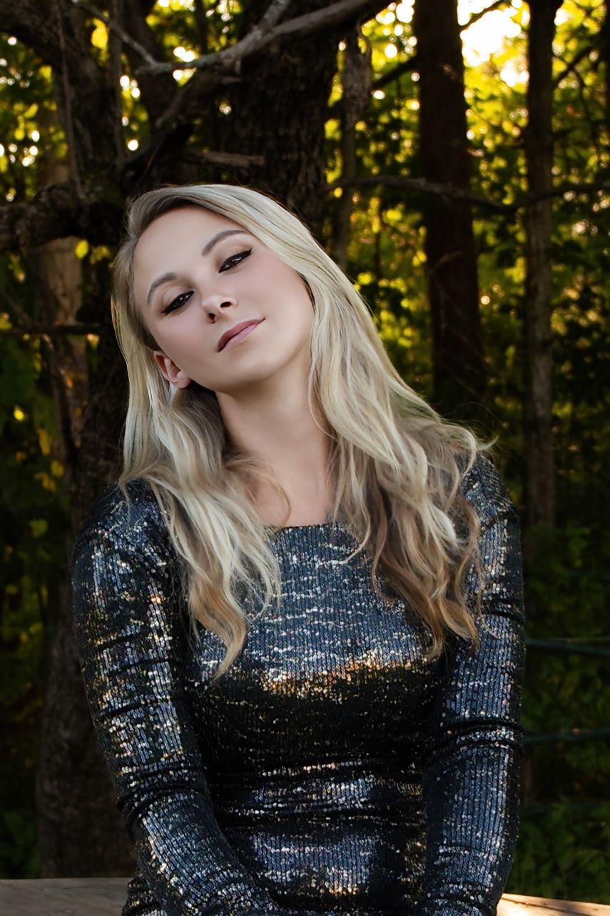 A figure in a sparkling sequined dress poses dramatically against a forest backdrop in golden evening light.