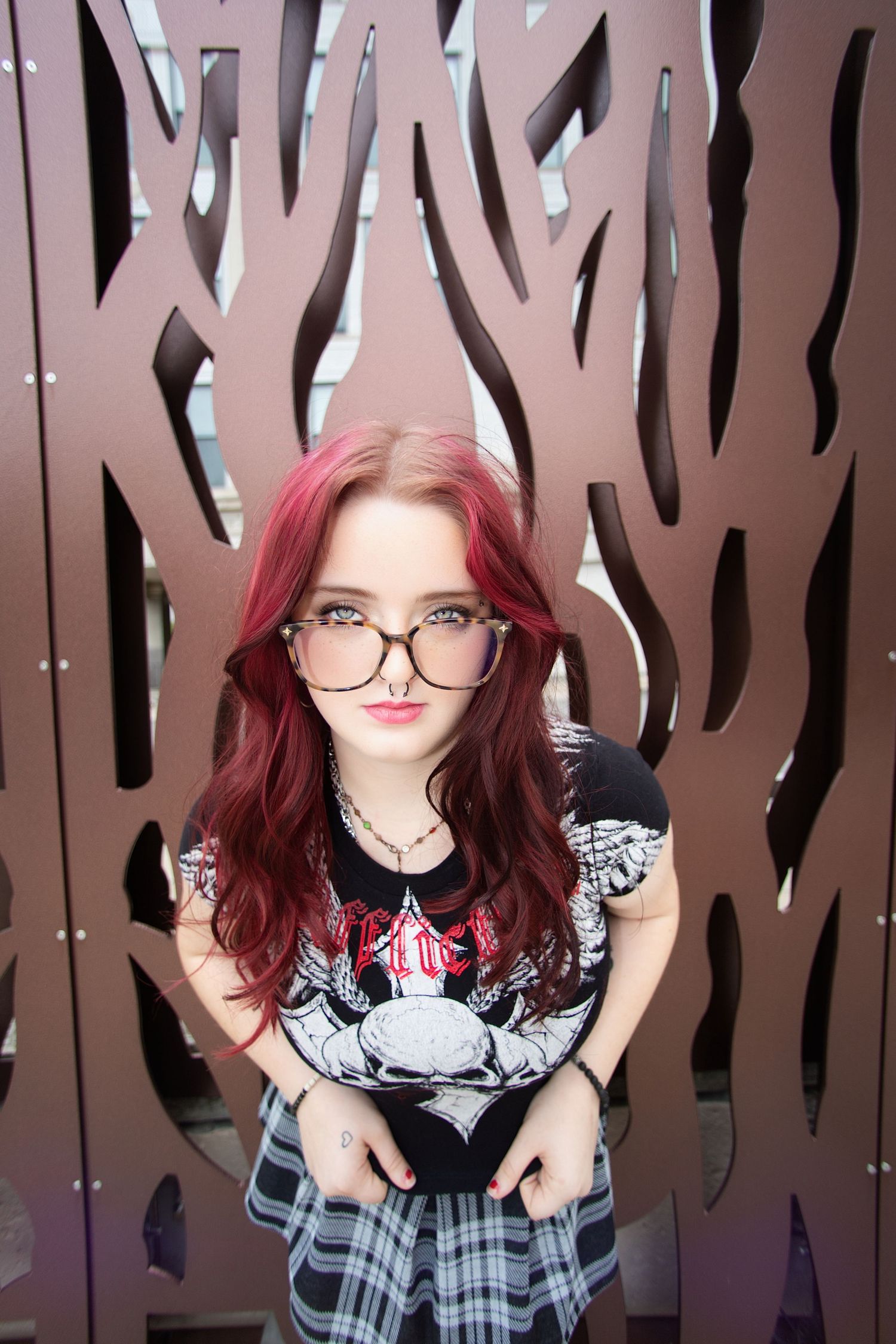 Young woman with red hair and glasses wearing a graphic tee and plaid skirt, posing in front of decorative metal wall.