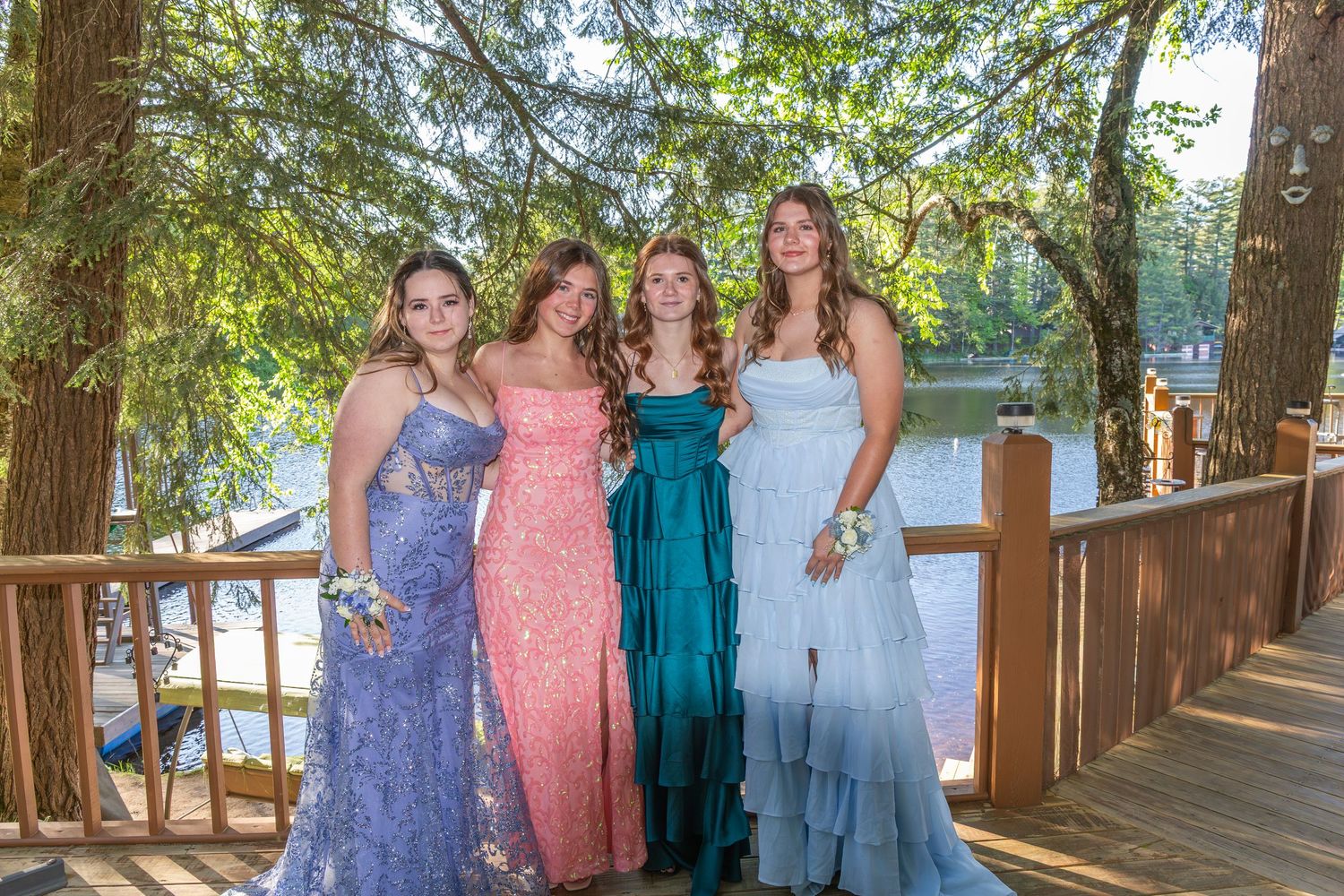 Four young women in elegant prom gowns pose together on a wooden deck near a lake surrounded by trees.